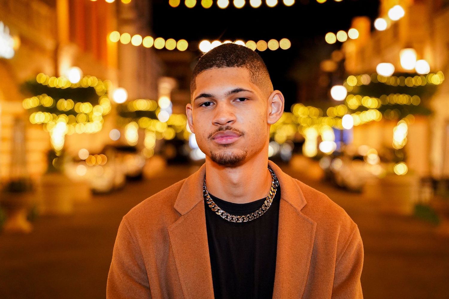 Person in camel coat and chain necklace poses under outdoor string lights at night.