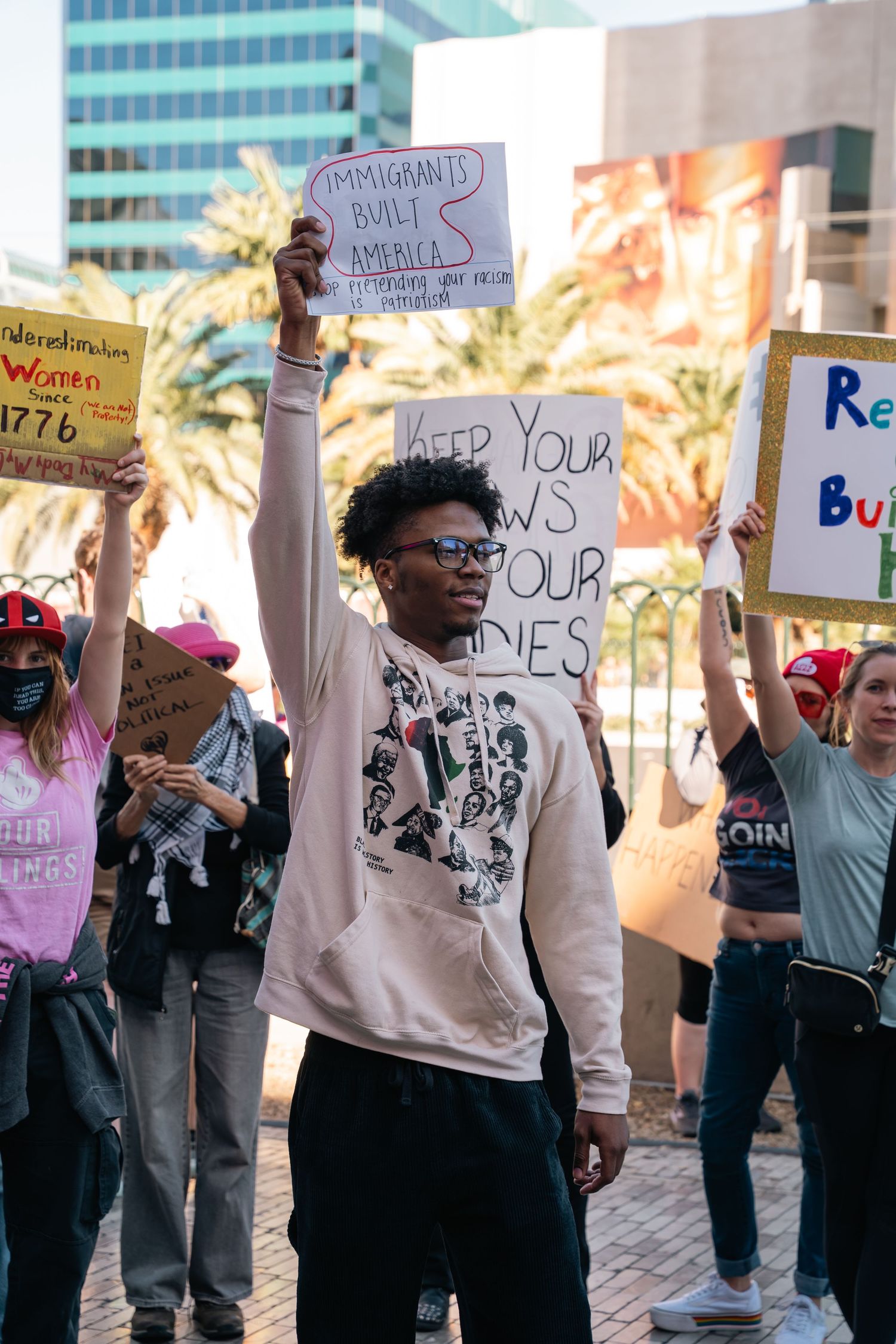 Demonstrators holding protest signs at a daytime rally in front of office buildings.