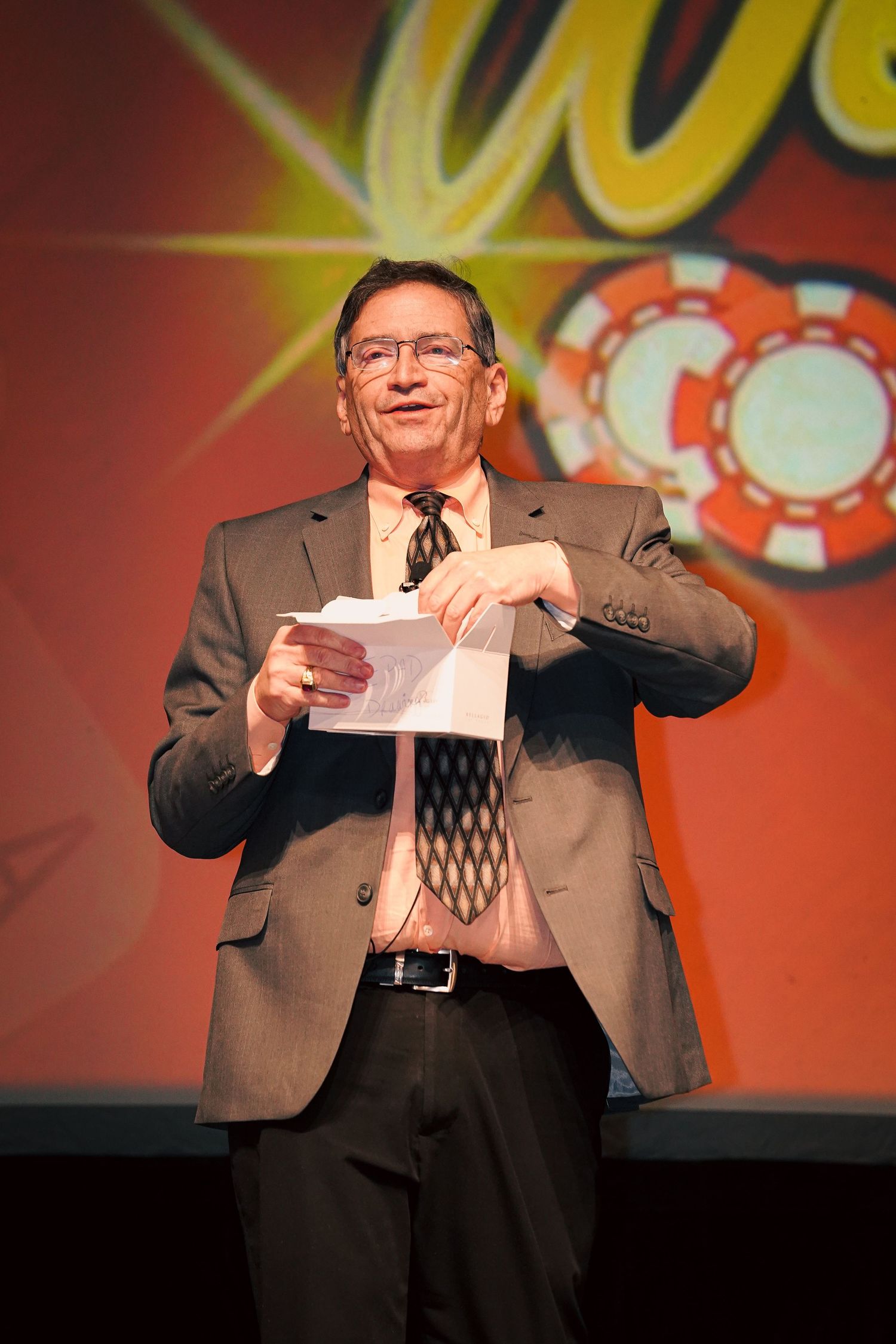 Speaker in suit smiles while holding papers on stage with casino-themed background.