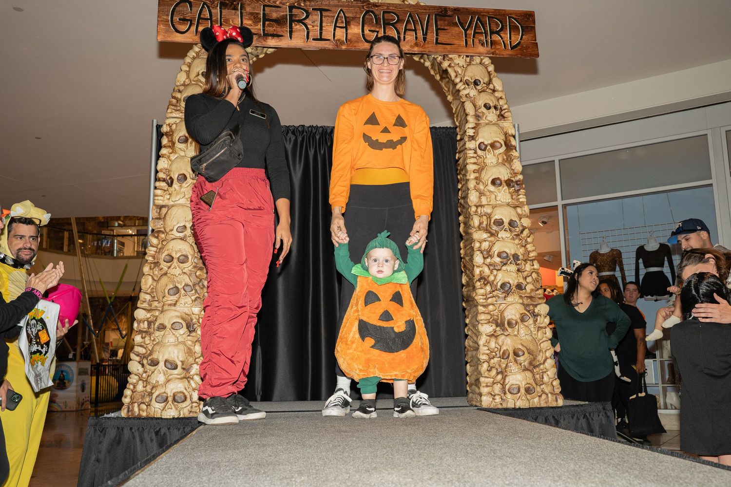 People in Halloween costumes stand under a decorated archway at a mall event.