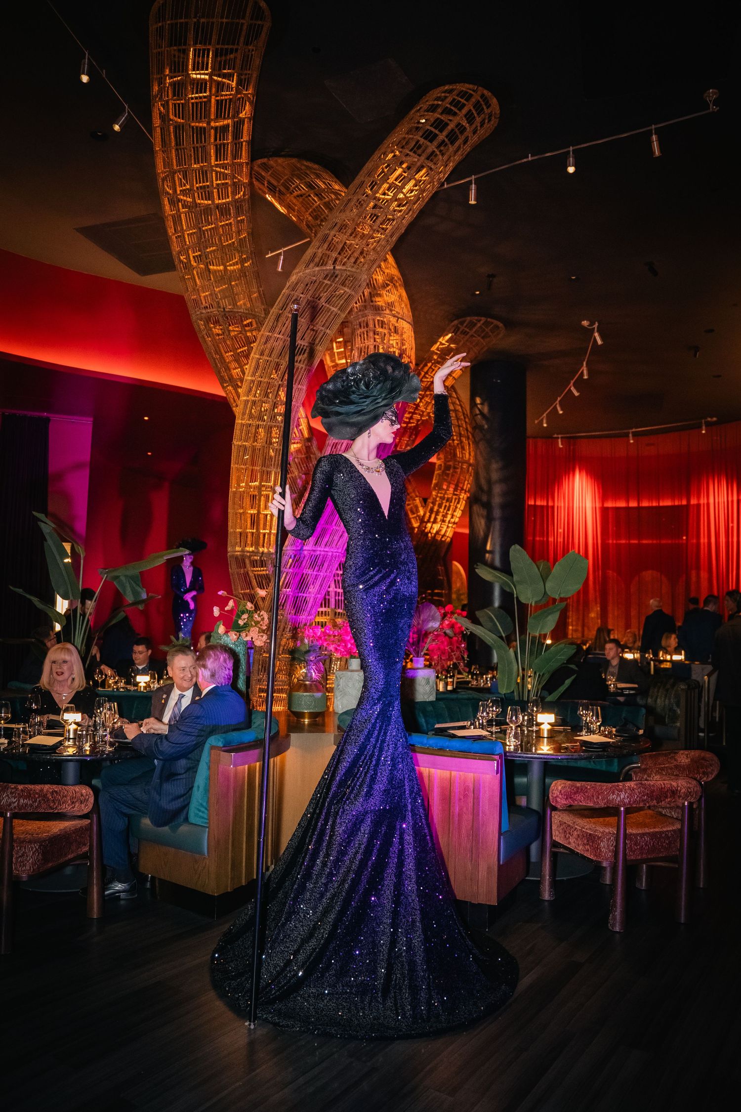 A performer in a navy sequined gown stands beneath a large woven sculpture in a dimly lit venue.