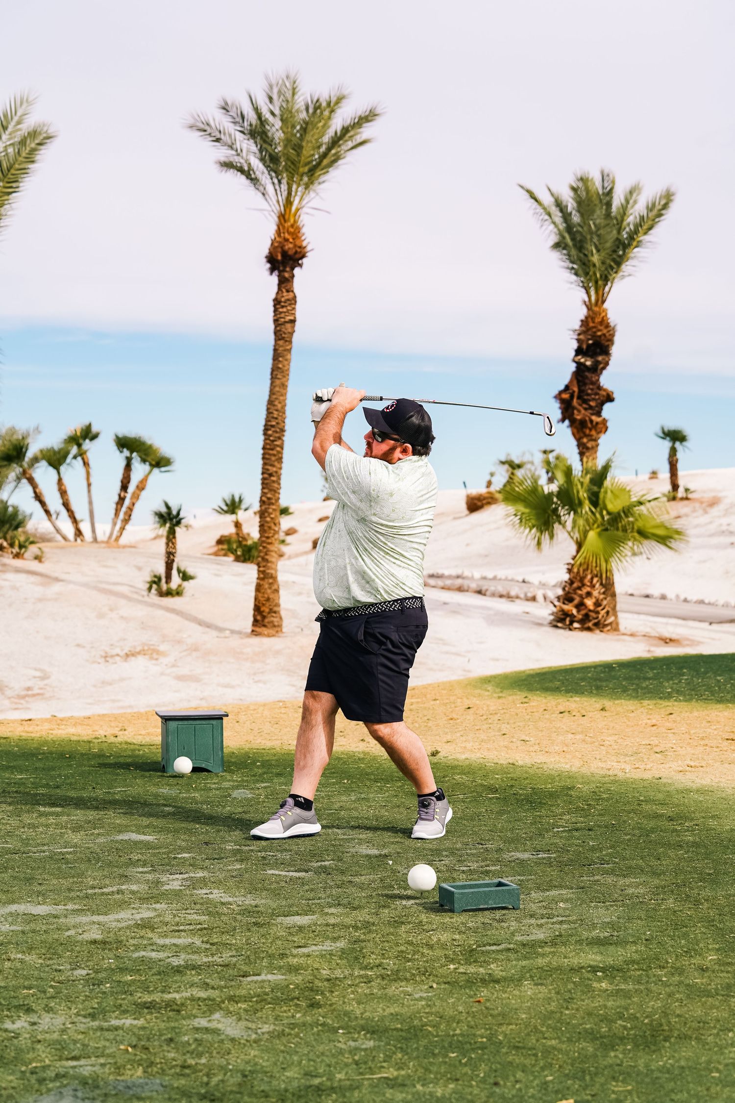 Golfer taking a swing at ball on tee with palm trees in desert landscape.