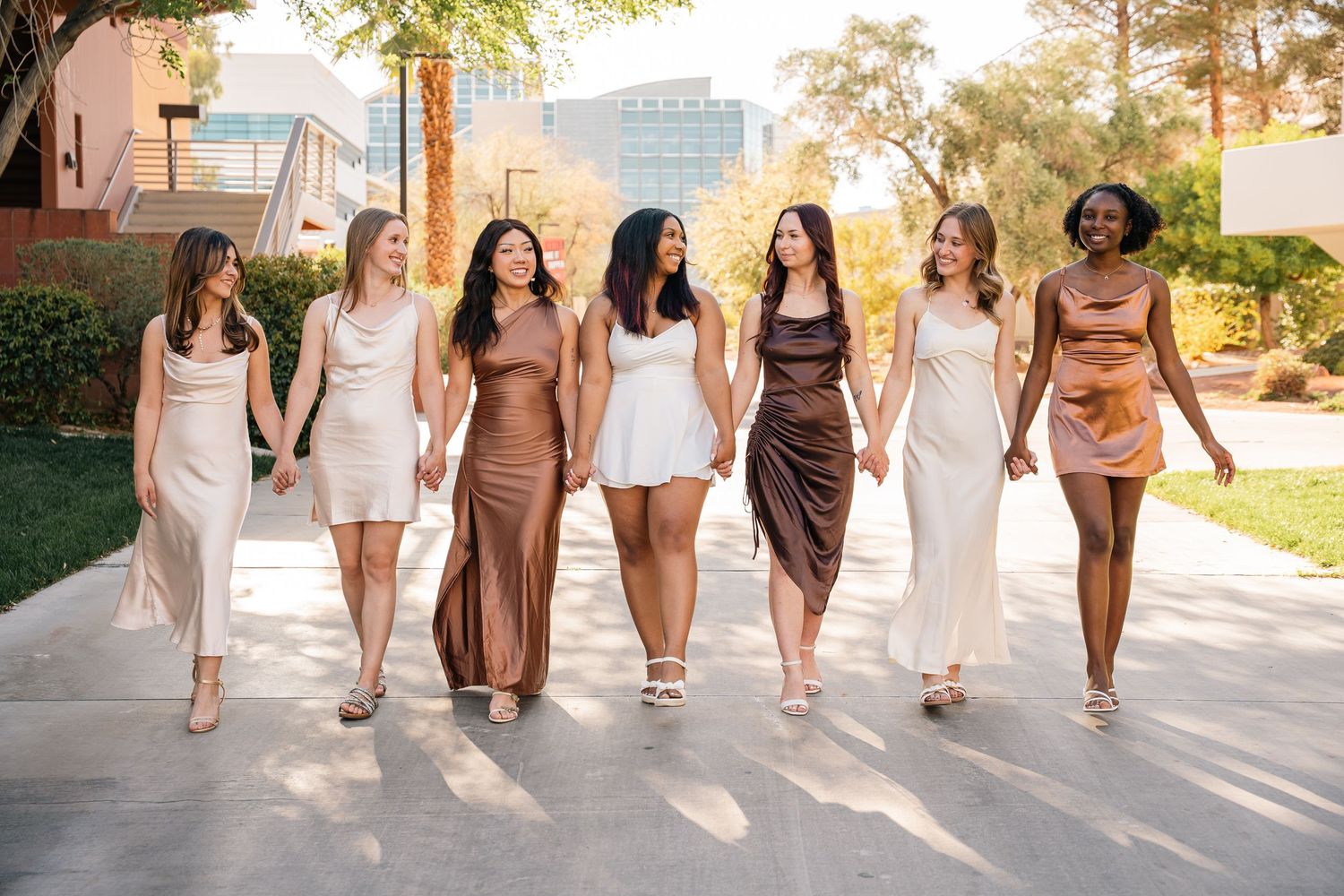 Group of women walking together wearing neutral toned dresses in sunlit outdoor setting.