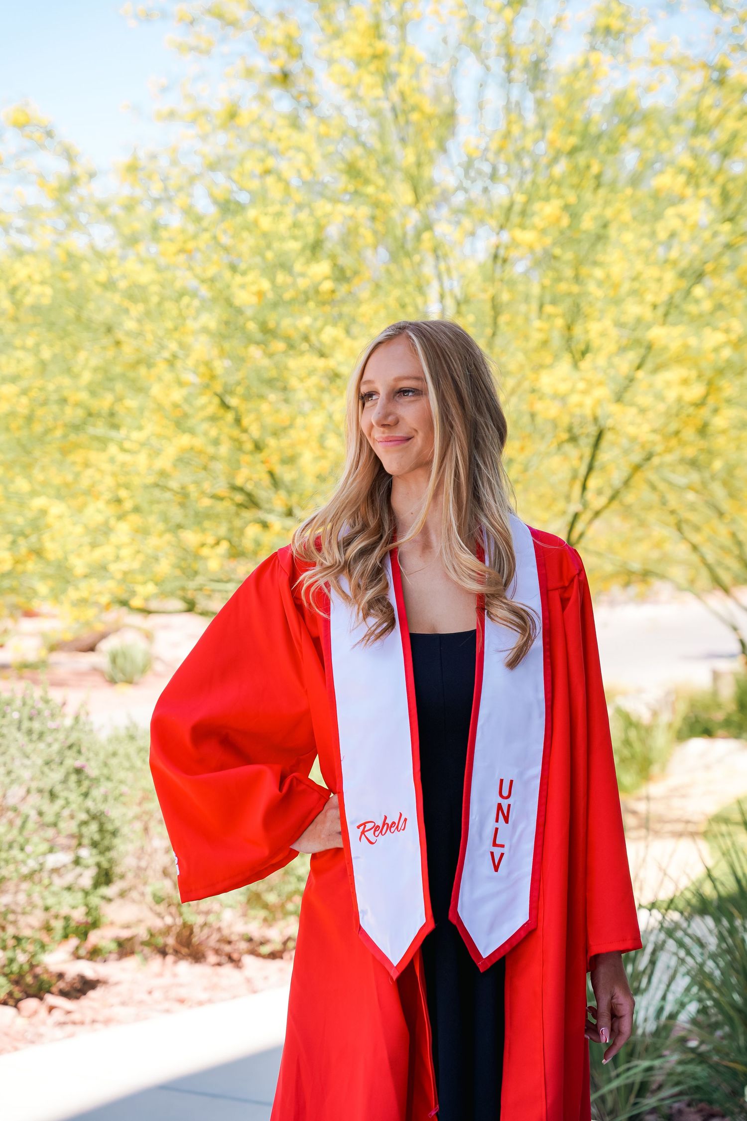 Graduate in red commencement robe poses against yellow flowering trees outdoors.