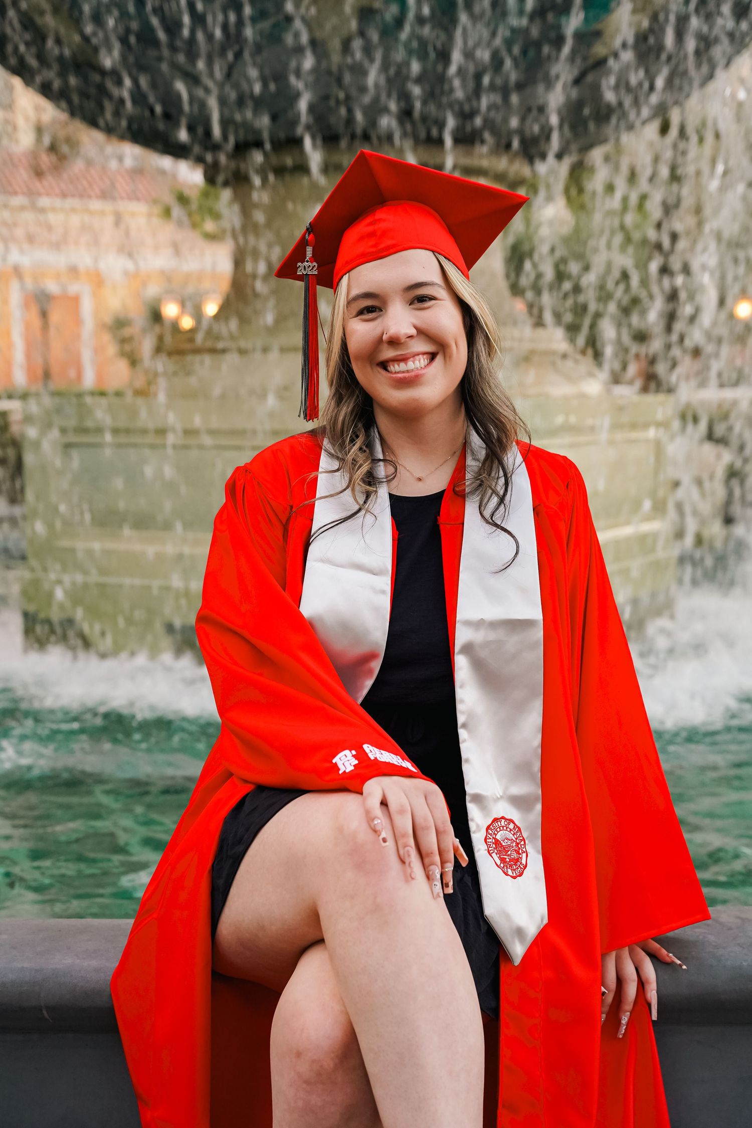 Graduate in red cap and gown poses by fountain in formal graduation attire.