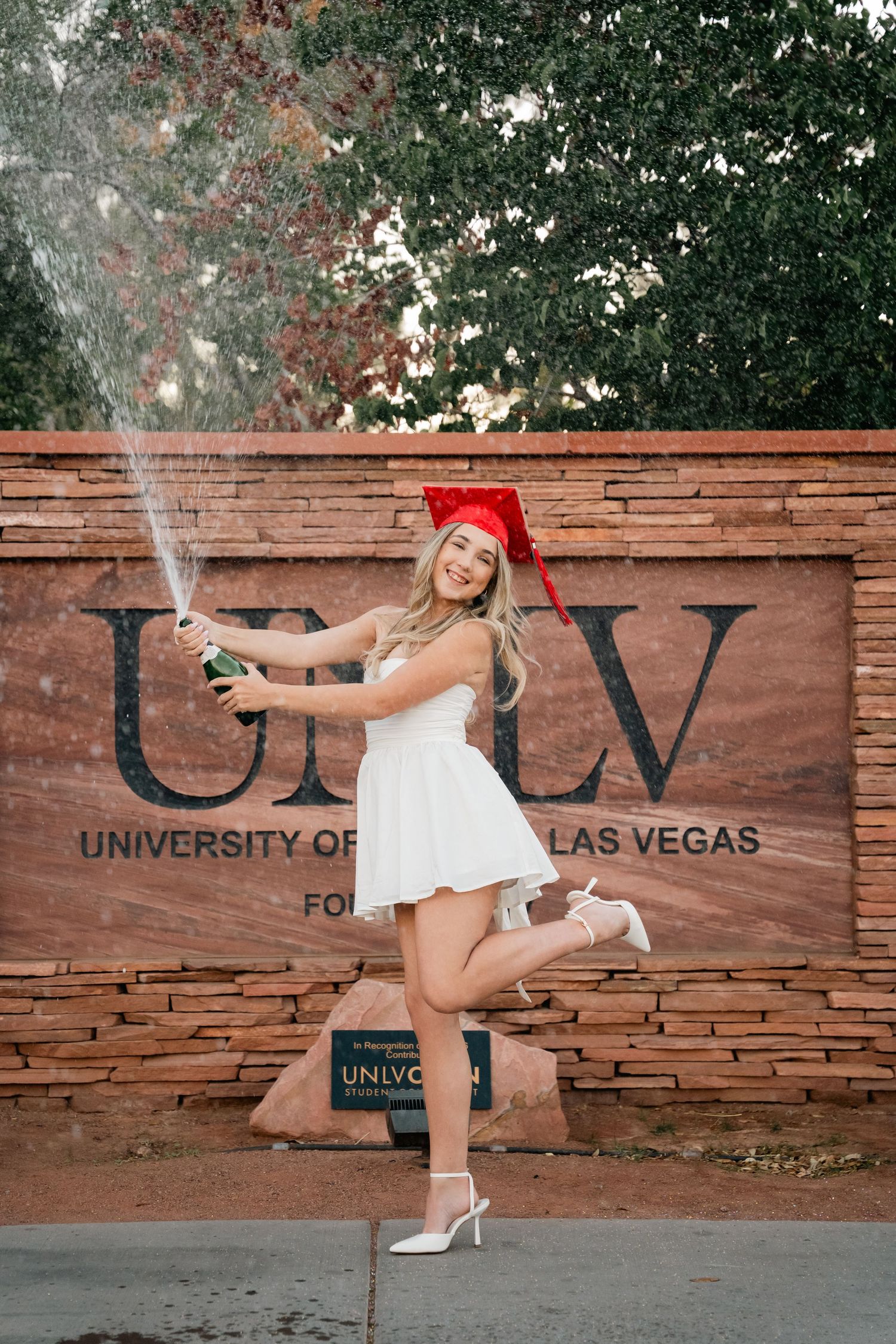 Graduate celebrating with champagne spray in front of UNLV sign wearing red cap and white dress.