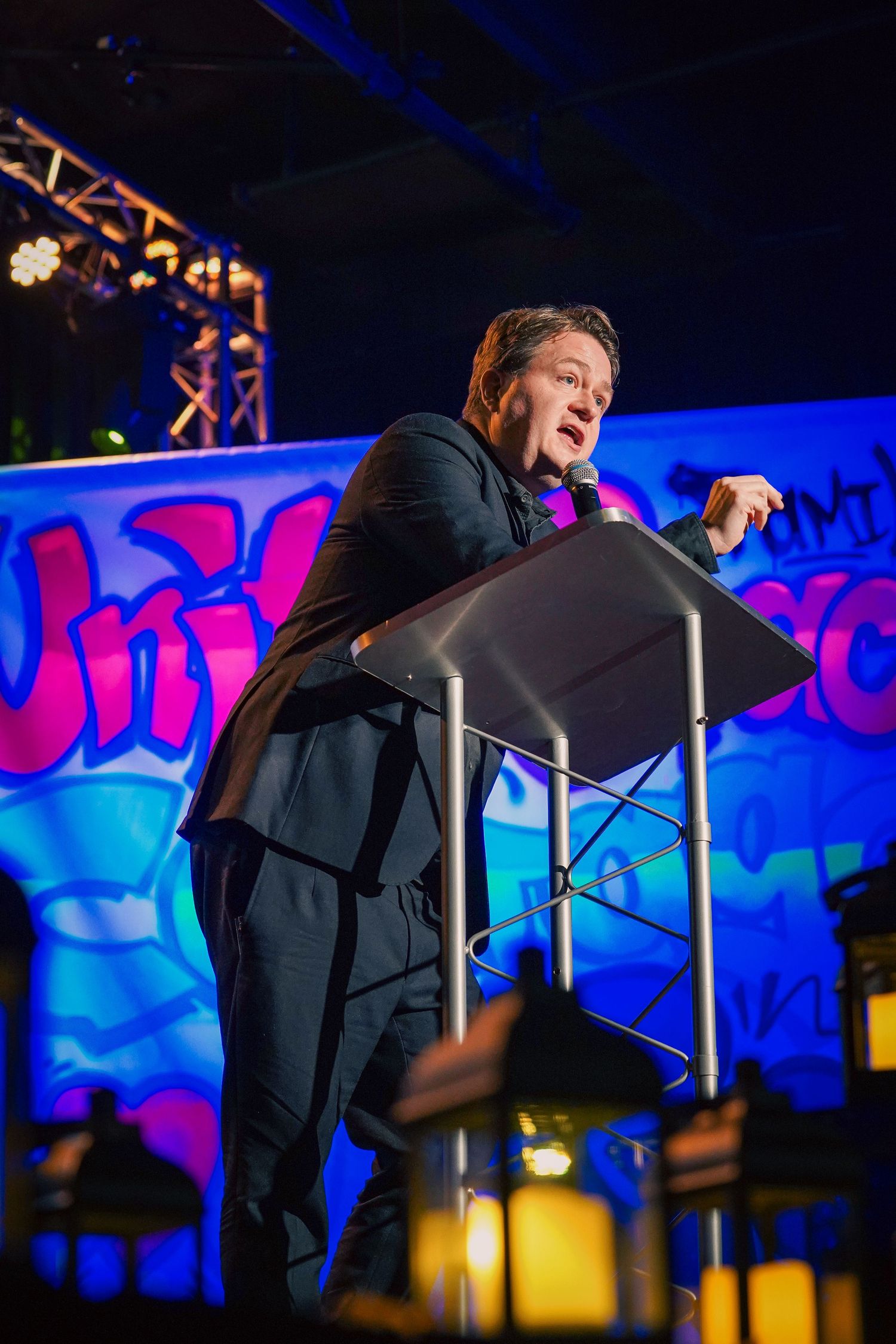 A speaker gestures energetically on stage with blue lighting and backdrop during a performance.