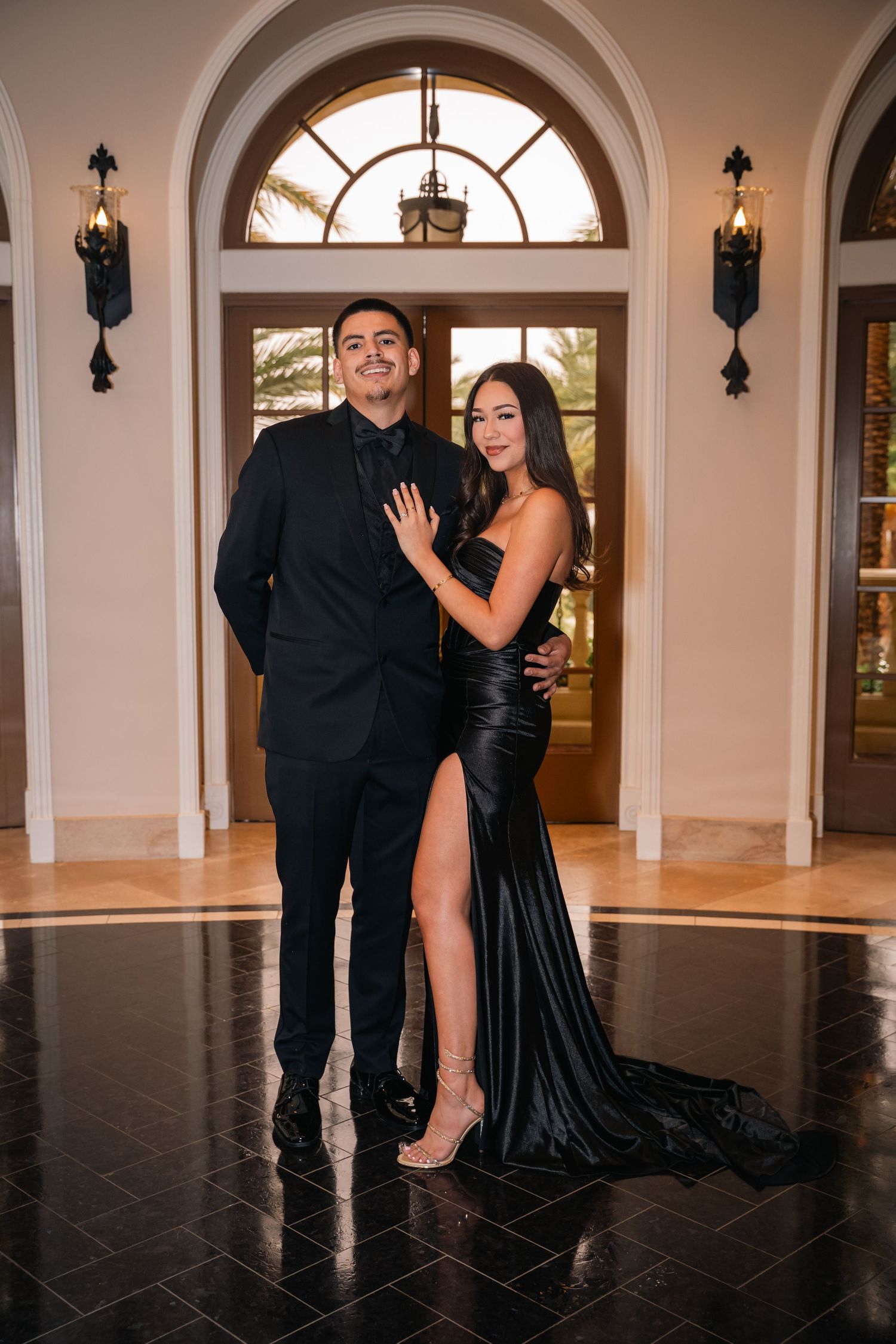 Elegant couple poses in formal attire in front of grand arched doorway with romantic lighting.