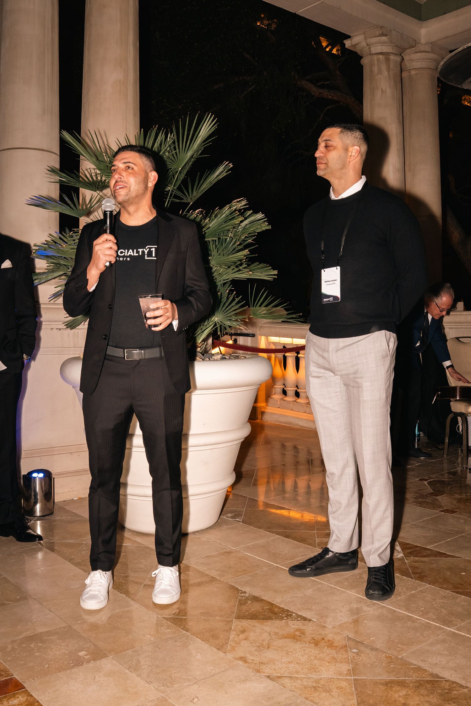Two attendees in smart casual attire stand near decorative planters at an evening business function.