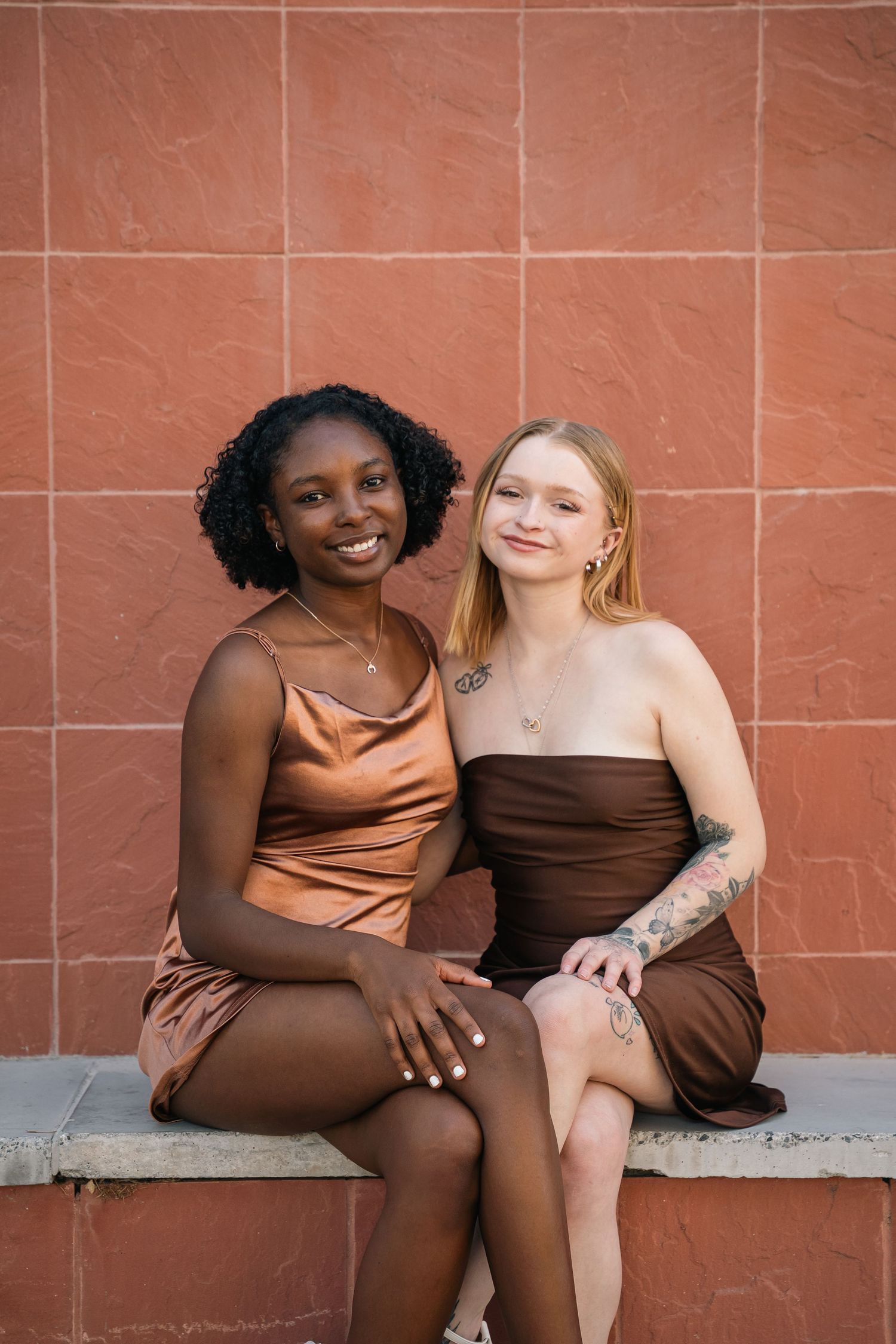 Two friends in brown and copper dresses sit together on a concrete bench.