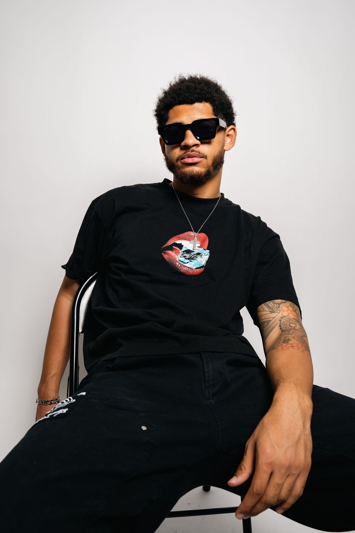 Monaco Media. Stylish person in black t-shirt with graphic print and sunglasses sitting on chair against white background.