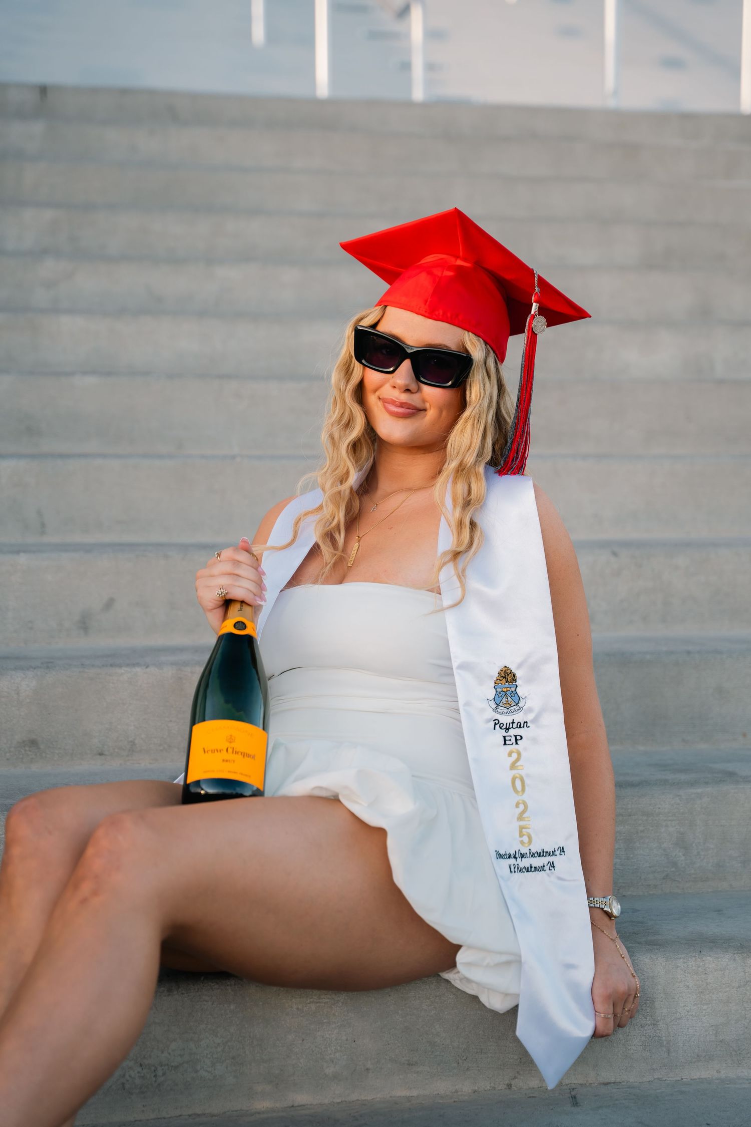 Graduate celebrates with champagne bottle while wearing red cap and white dress on concrete steps.