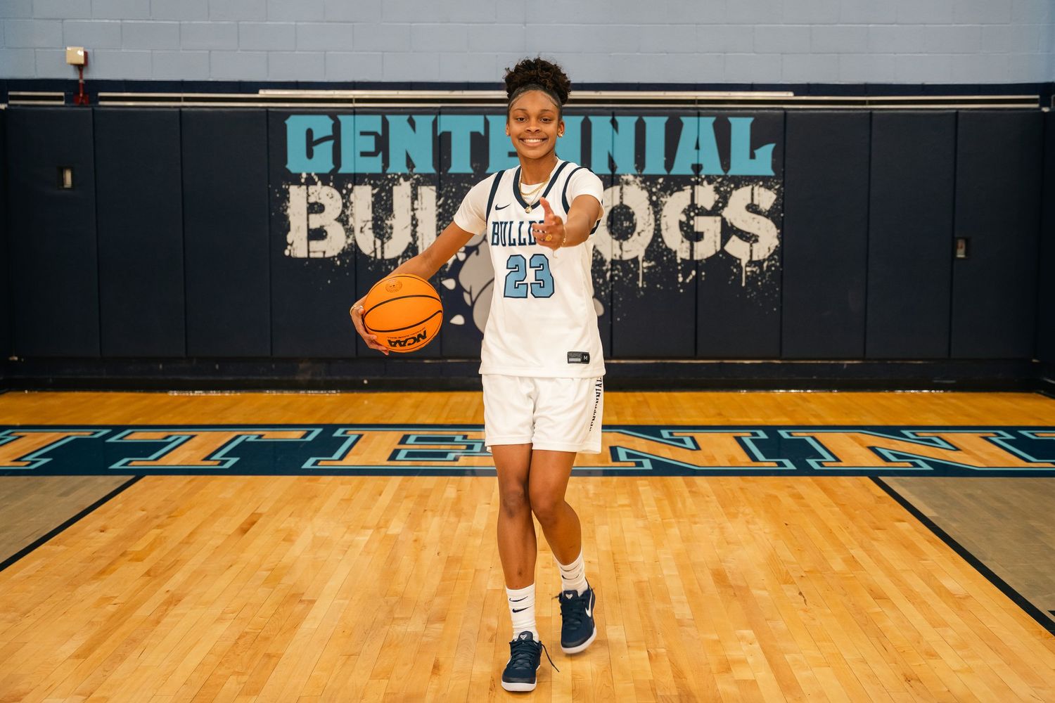Basketball player in white jersey number 23 poses with ball on court with Centennial Bulldogs logo.