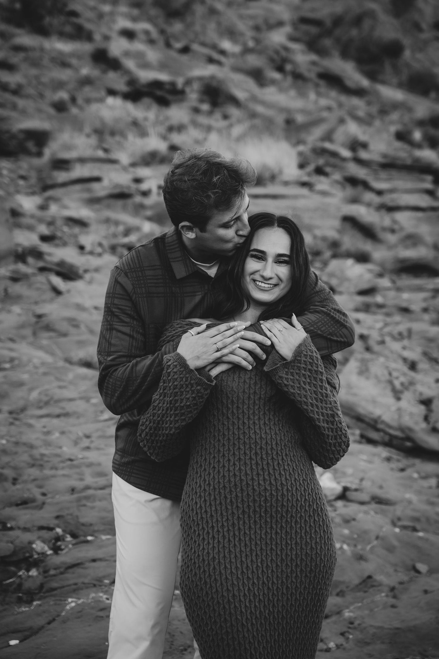 A loving couple embracing in black and white photography with desert rocks in the background.