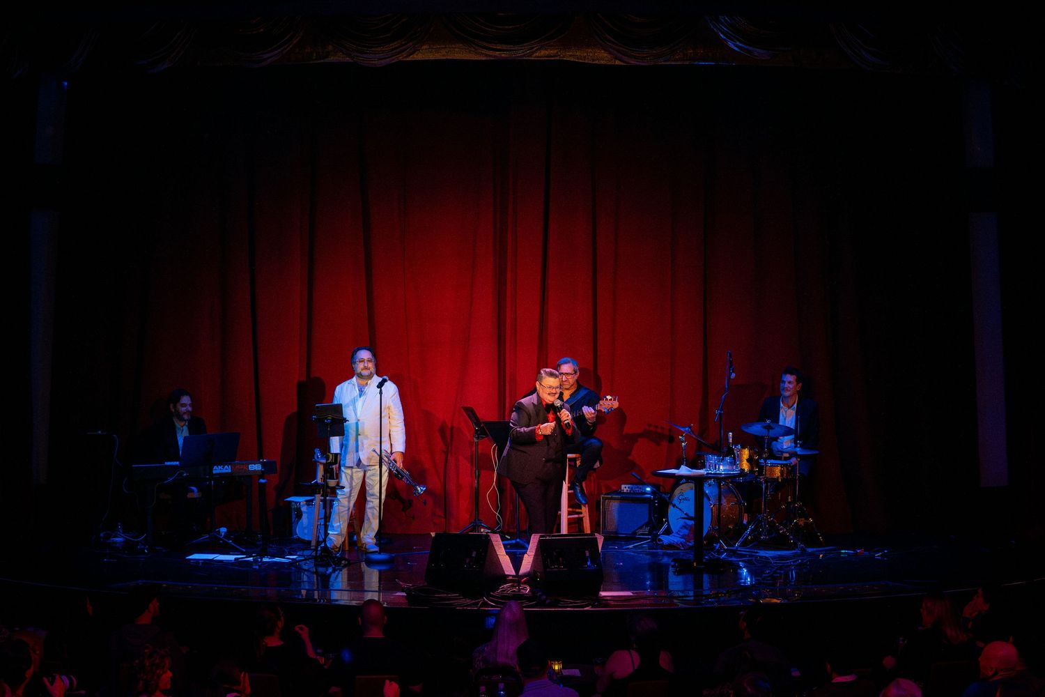 Musicians perform on stage with red curtain backdrop and theatrical lighting.