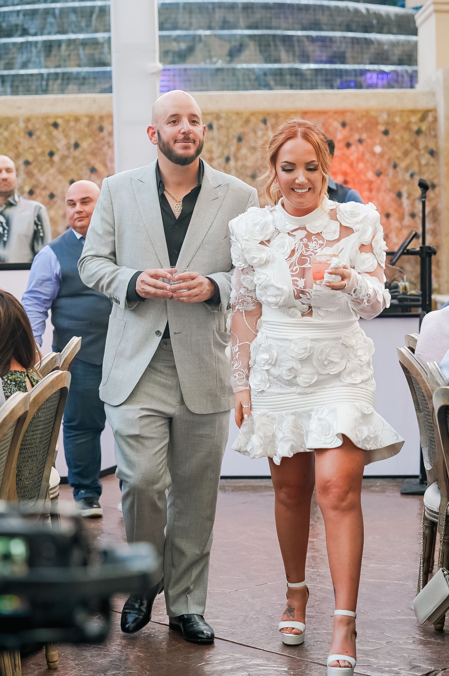 Couple walking together at reception, with person wearing light gray suit and white floral dress.
