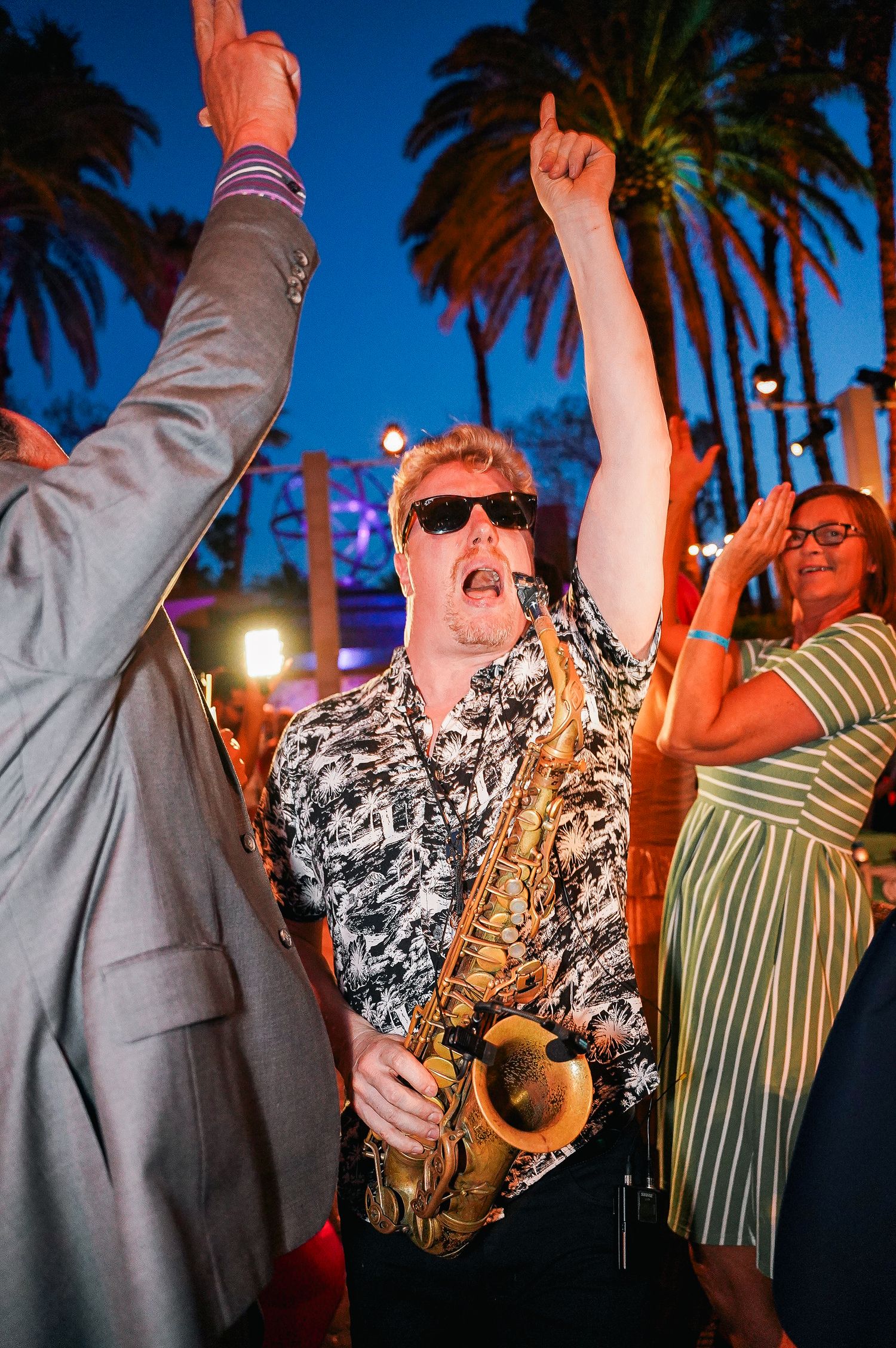 Outdoor evening celebration with palm trees and party guests raising their hands.