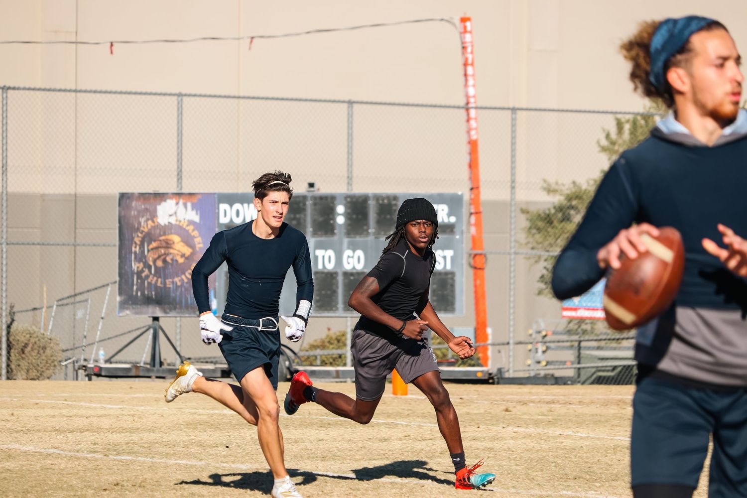 Athletes running football drills during practice session on dirt field.