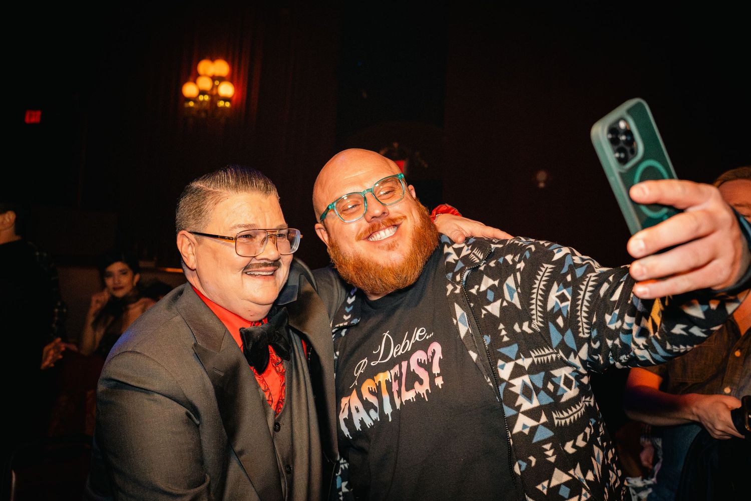 Two people take a selfie together at a dimly lit event venue with warm lighting in the background.