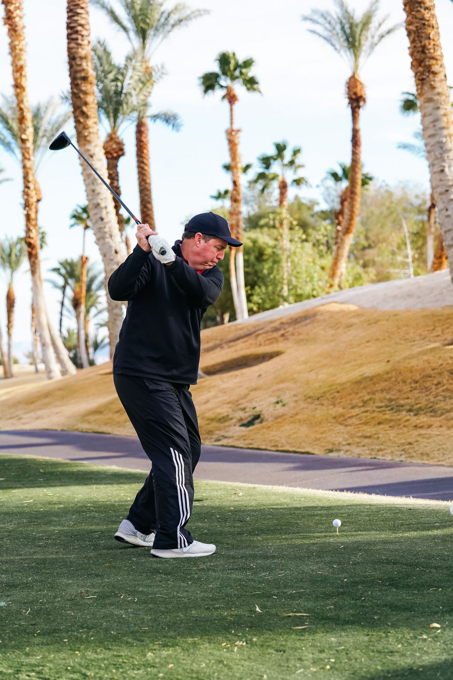 Golfer in dark athletic wear completing golf swing follow-through on desert course with palm trees.