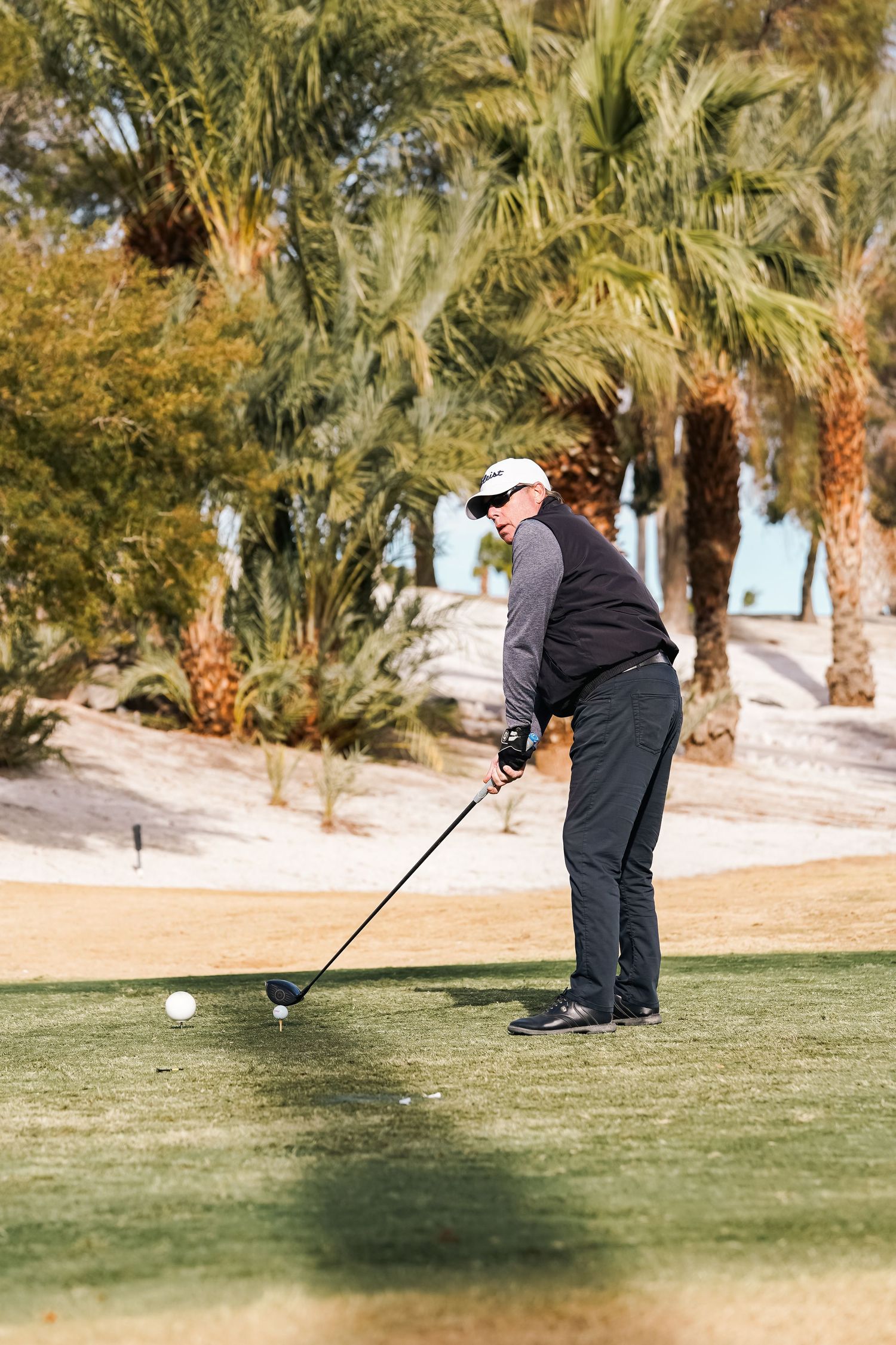 Golfer in dark clothing addressing the ball on a desert course with palm trees.