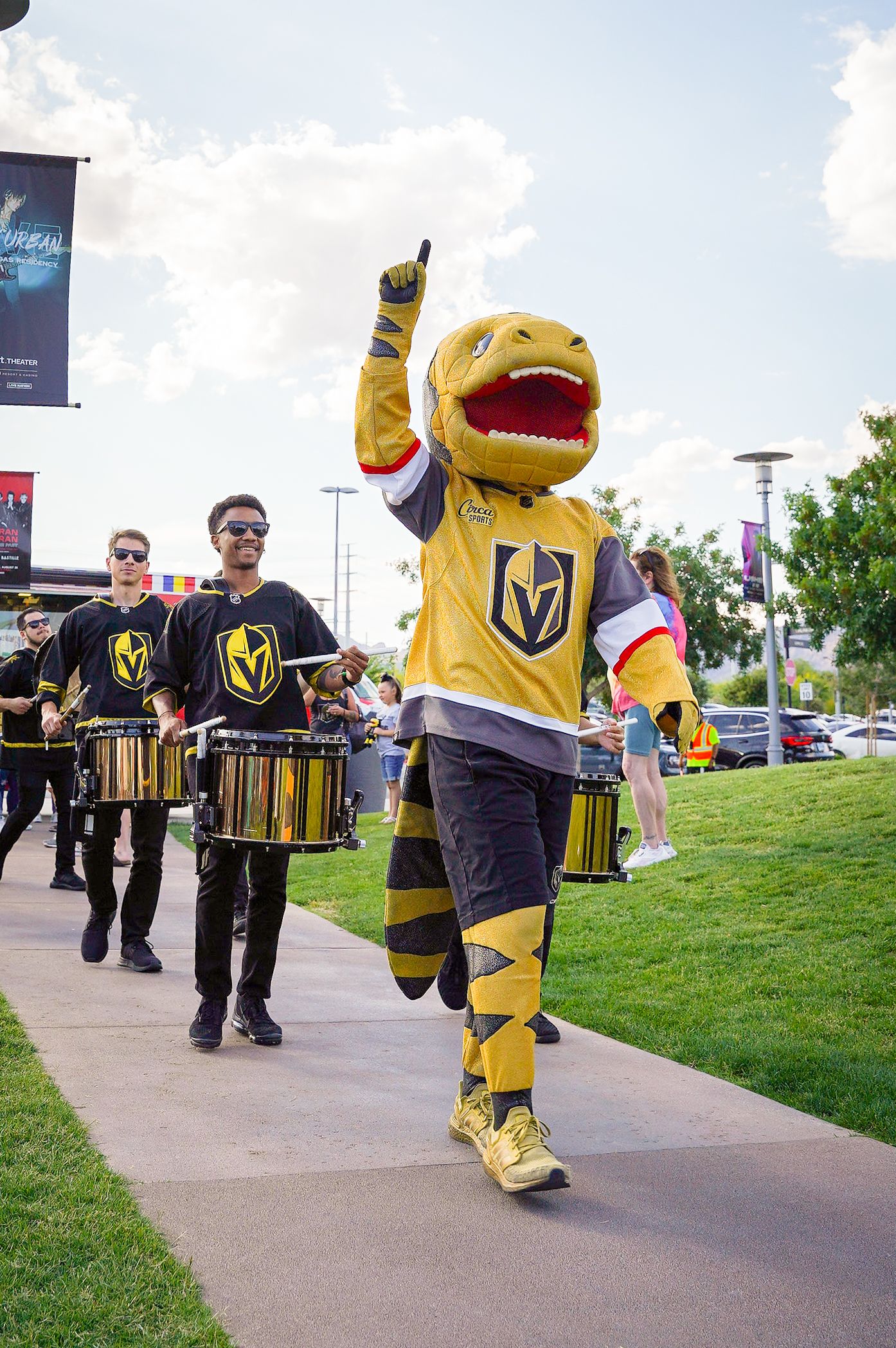 Vegas Golden Knights mascot marches with drum line performers on sidewalk at outdoor event.