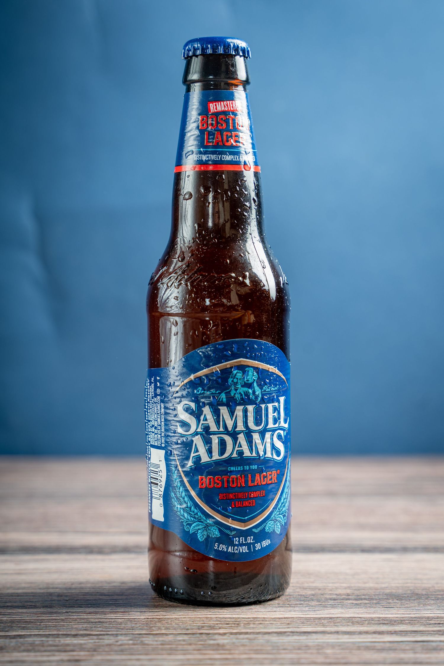 Cold bottle of Samuel Adams beer on wooden surface against blue background.