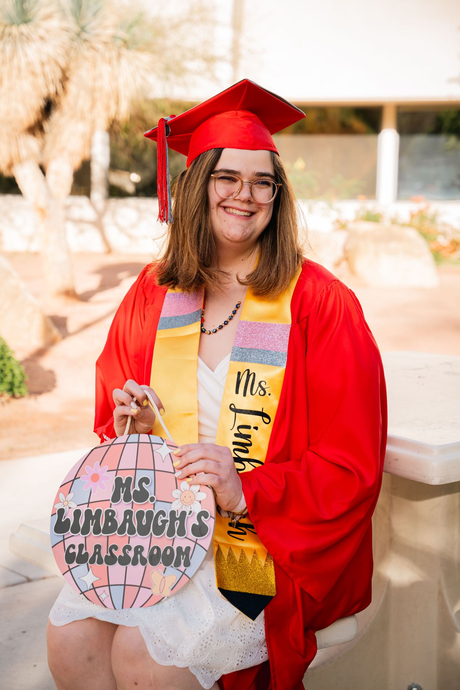 Graduate in red cap and gown holding classroom sign and decorated tumbler.