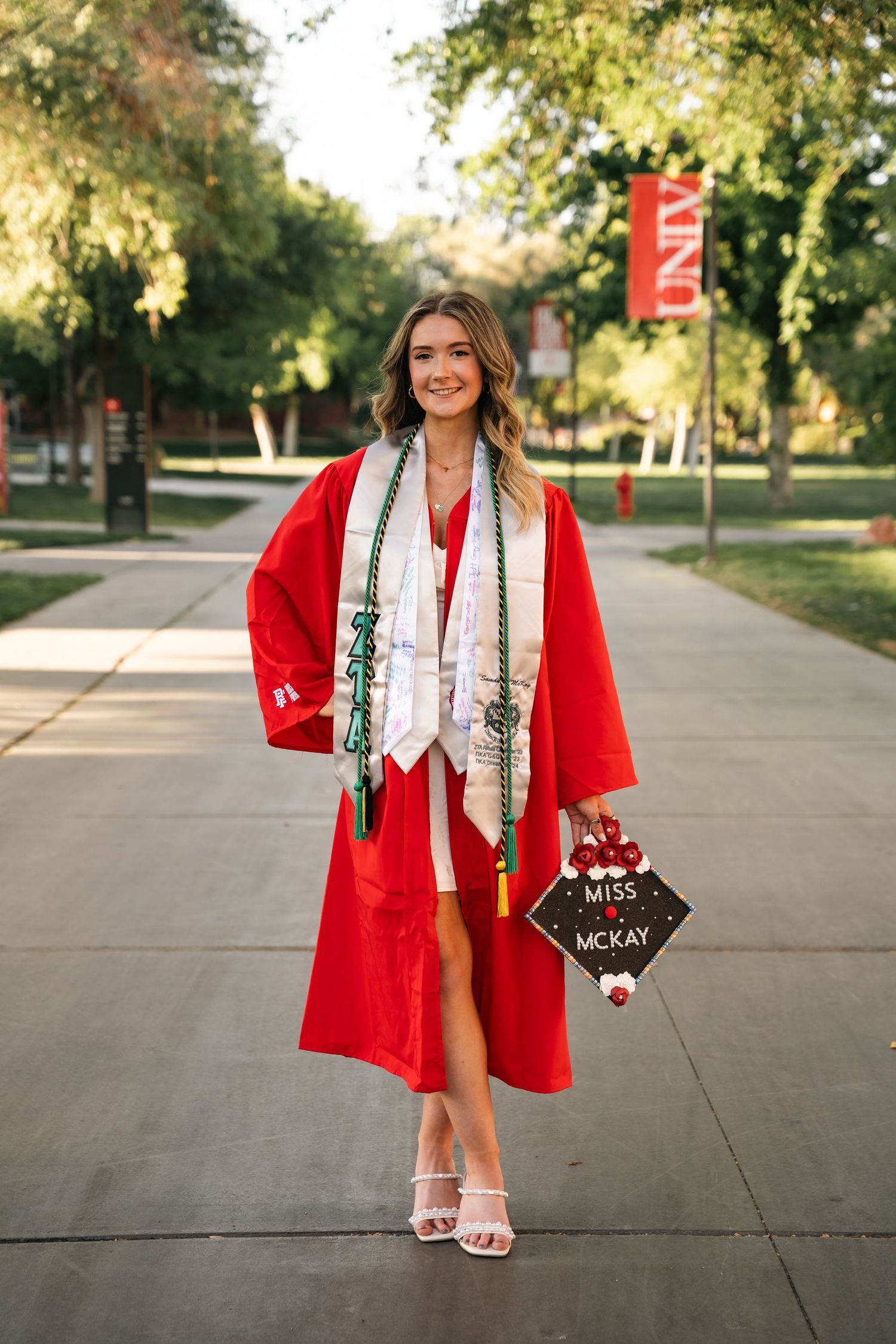 Graduate in red gown and white dress poses with decorated cap on university campus pathway.