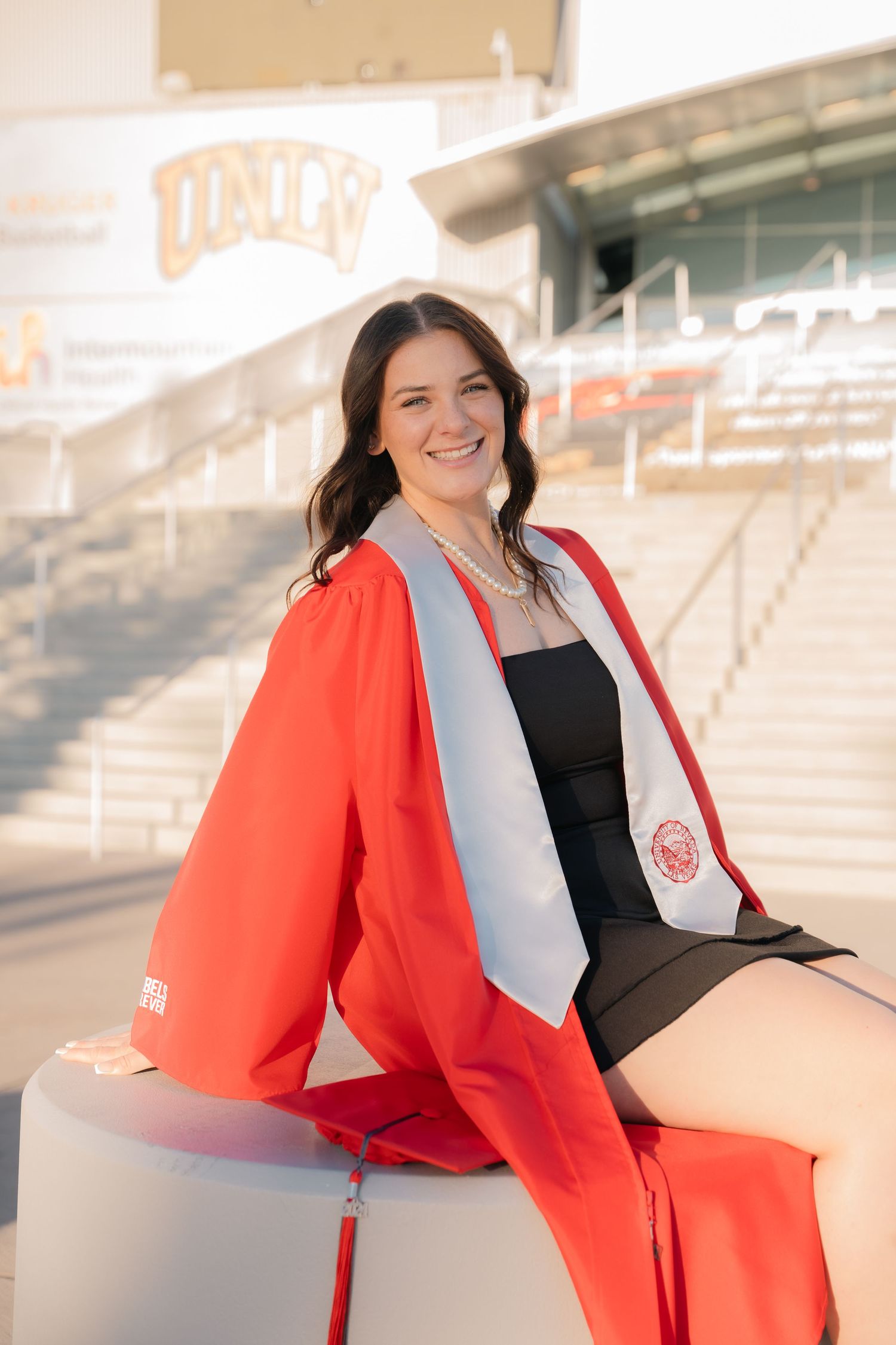 Graduate in red graduation gown poses near stadium seating.