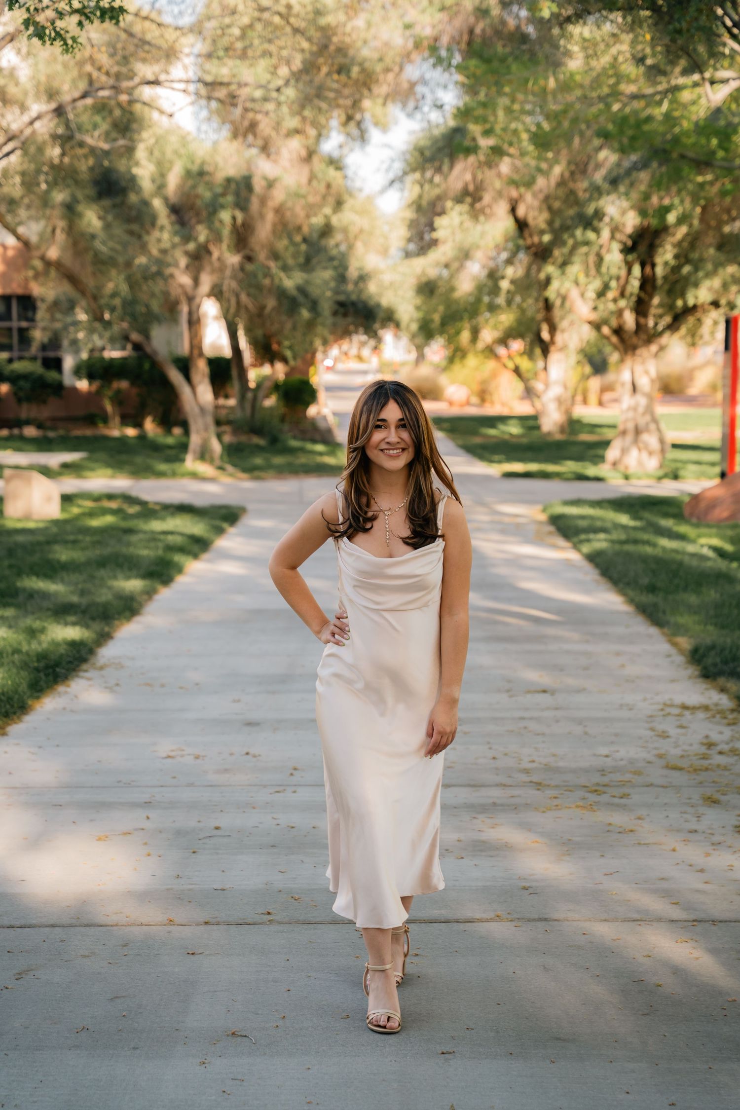 Woman in cream-colored dress standing on concrete path with trees in background.