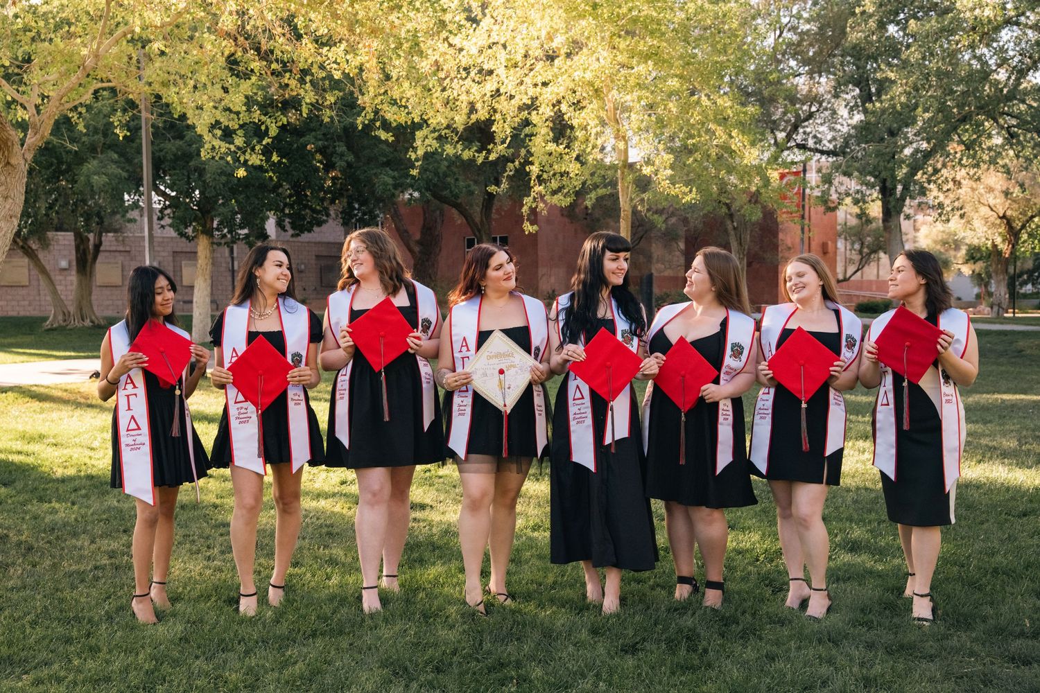 Group in black dresses with red stoles pose for graduation photos on a sunny campus lawn.