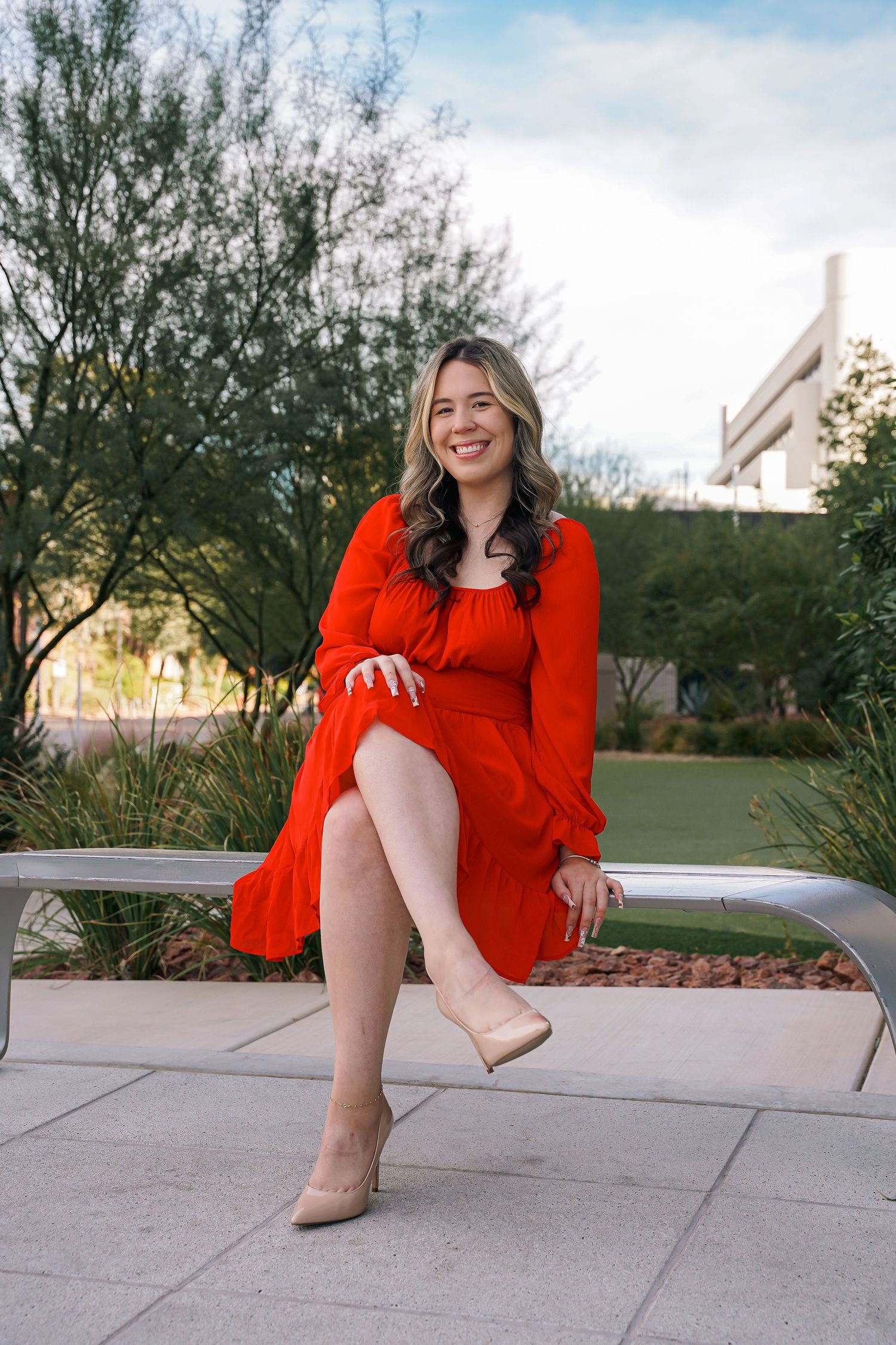 Graduate in red dress sits casually on bench during outdoor photo session.