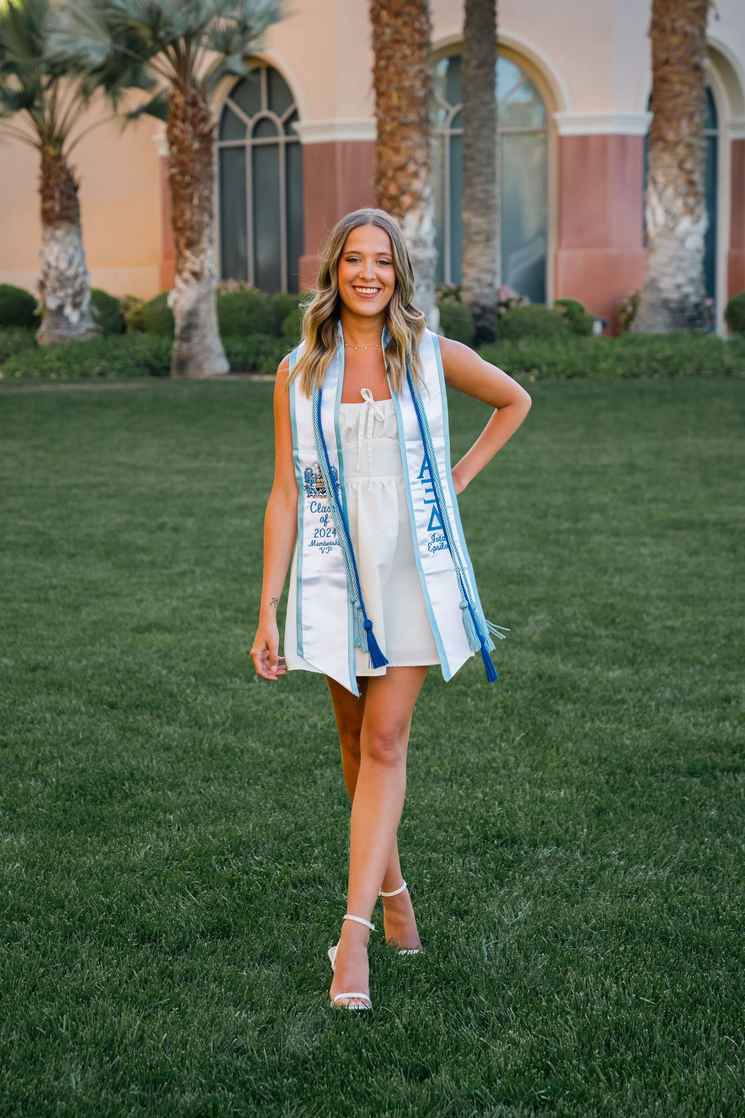 Graduate in white dress and blue sash poses cheerfully on lawn with Spanish colonial architecture.