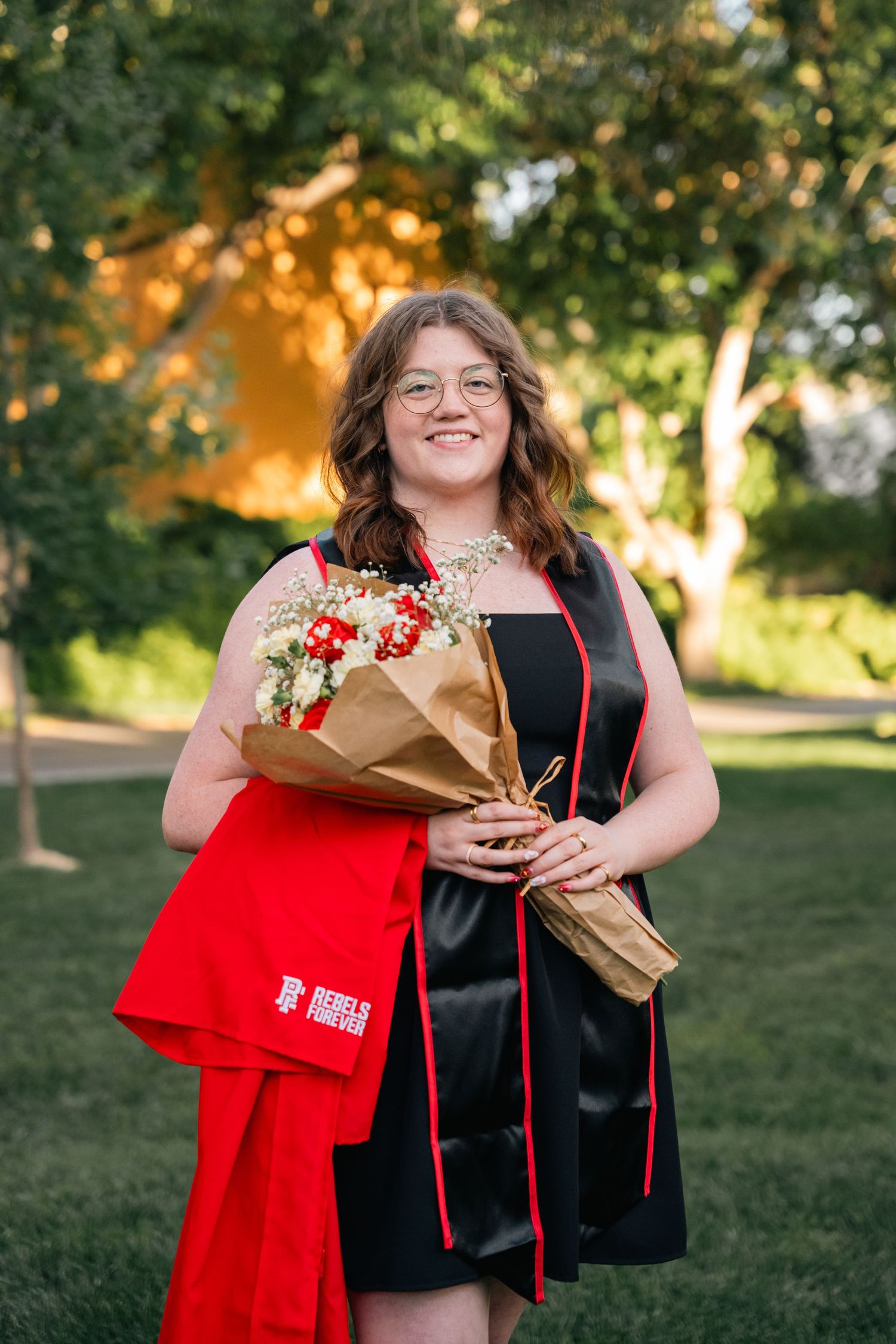 Graduate in black cap and gown holds bouquet of red roses while smiling outdoors.