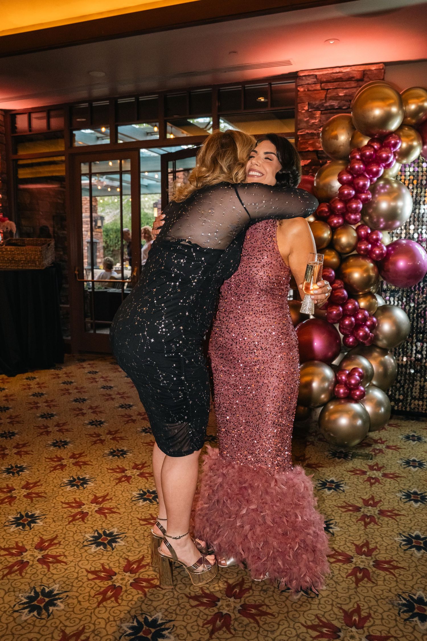 Two guests in evening wear embrace at a party with a balloon arch backdrop.