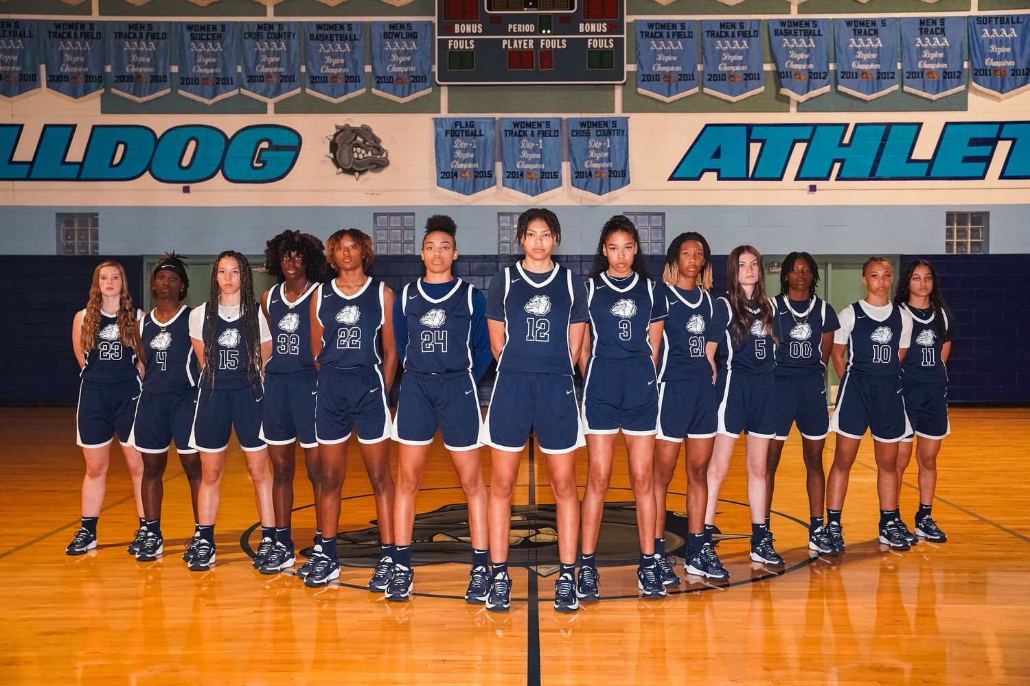 Basketball team in navy uniforms stands in v-formation on court under Bulldog Athletics banner.