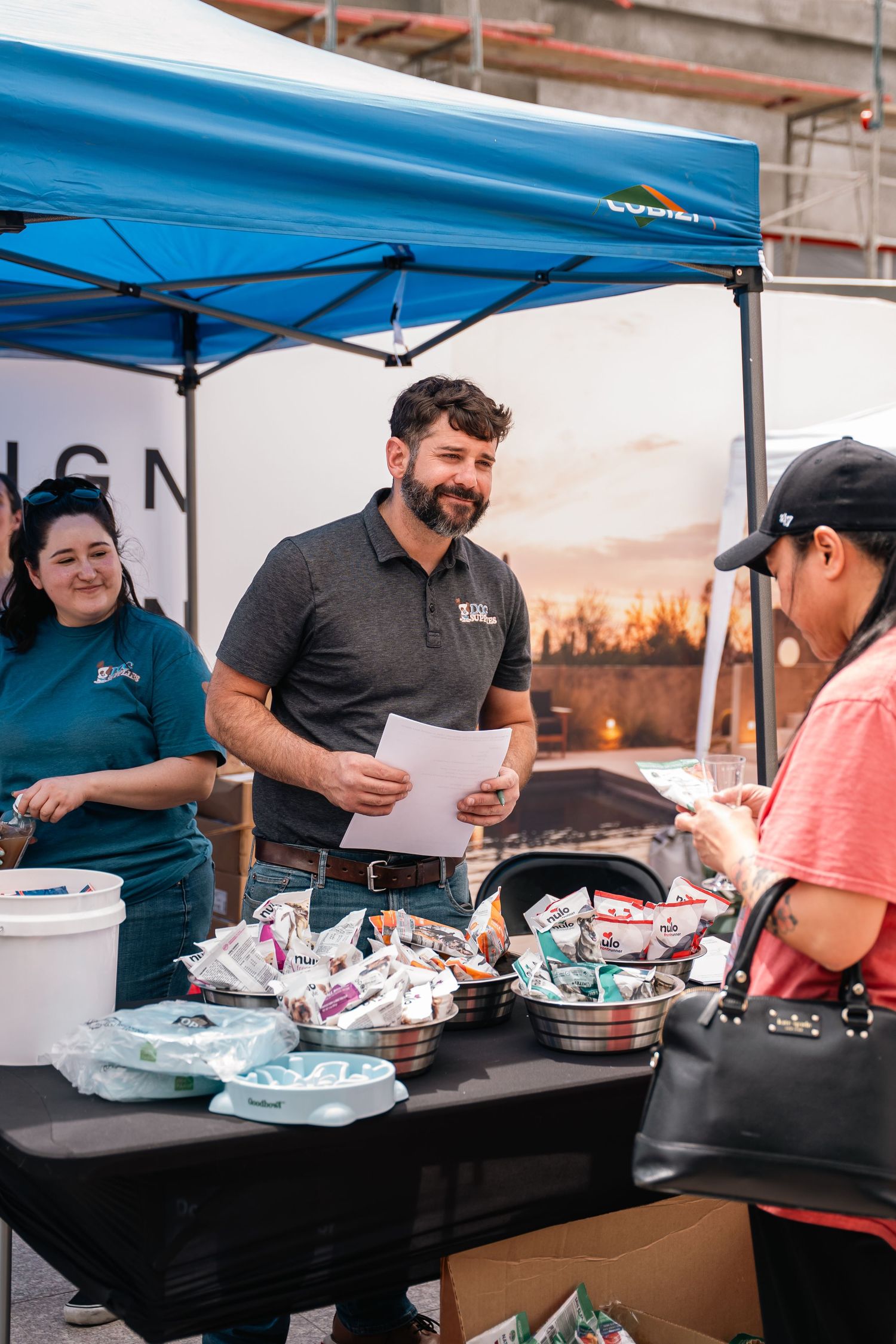 Staff serve food at an outdoor event under a blue tent with buffet trays on a black table.
