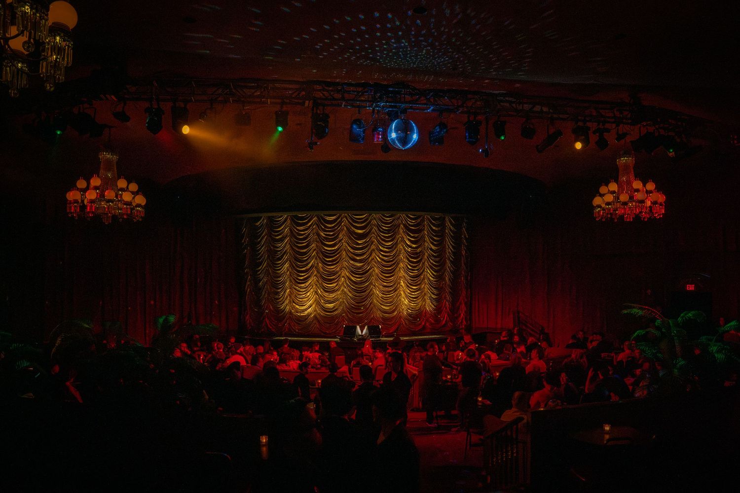 Theater audience awaits performance in dimly lit venue with ornate gold curtain and red lighting.