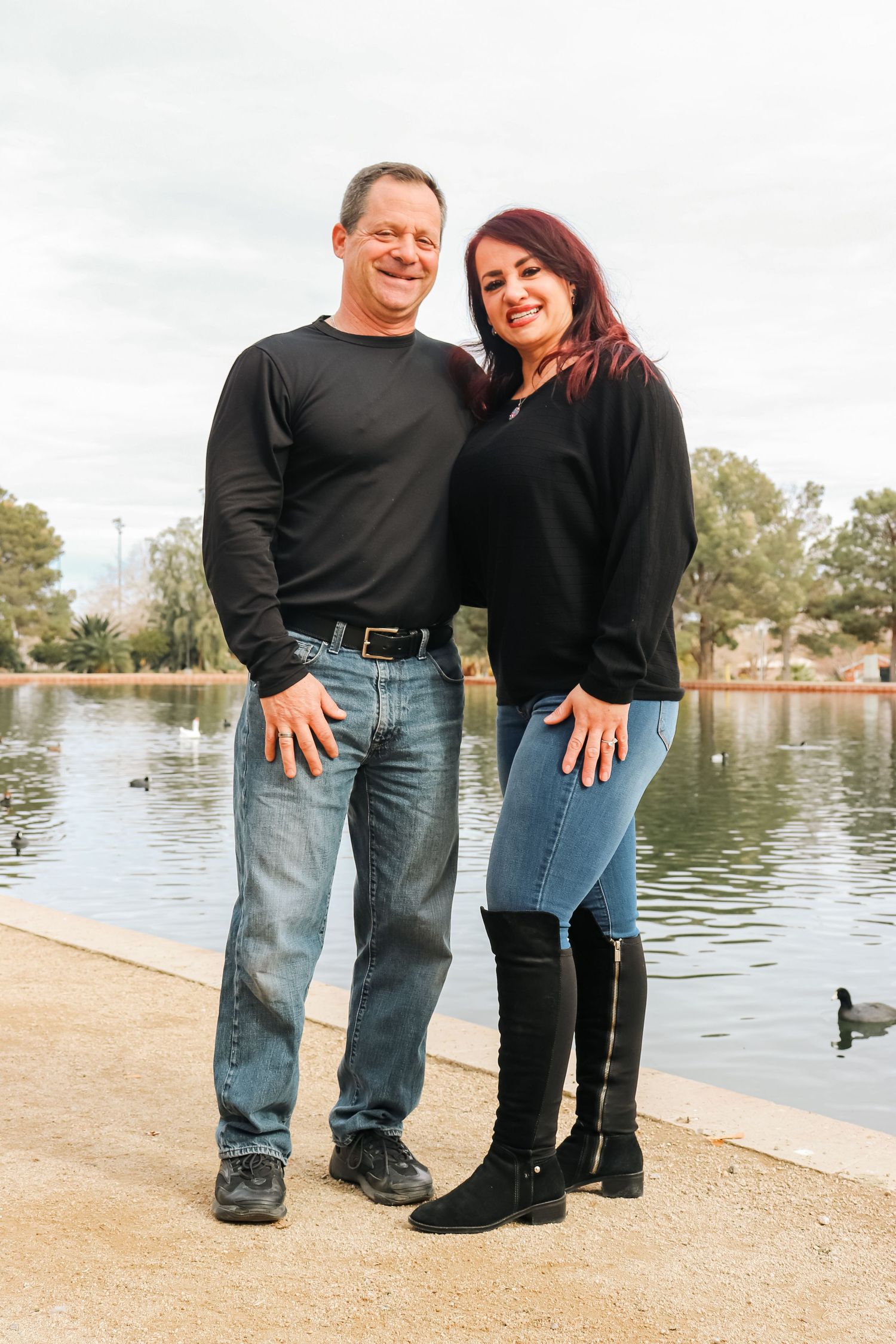 Couple in matching black sweaters pose together by a lake with ducks swimming in background.