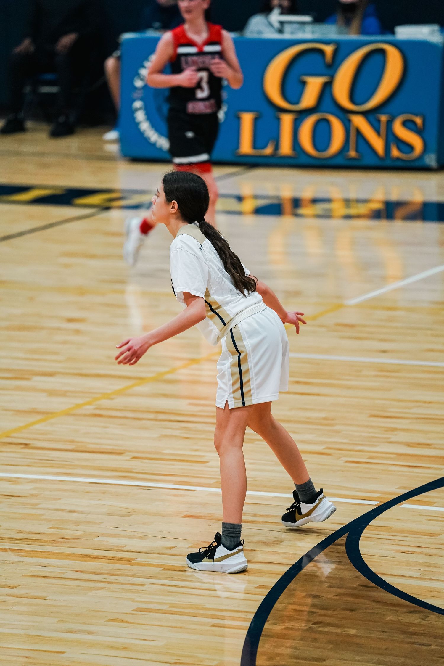 Basketball player in white uniform stands ready on court with 'Go Lions' banner visible.