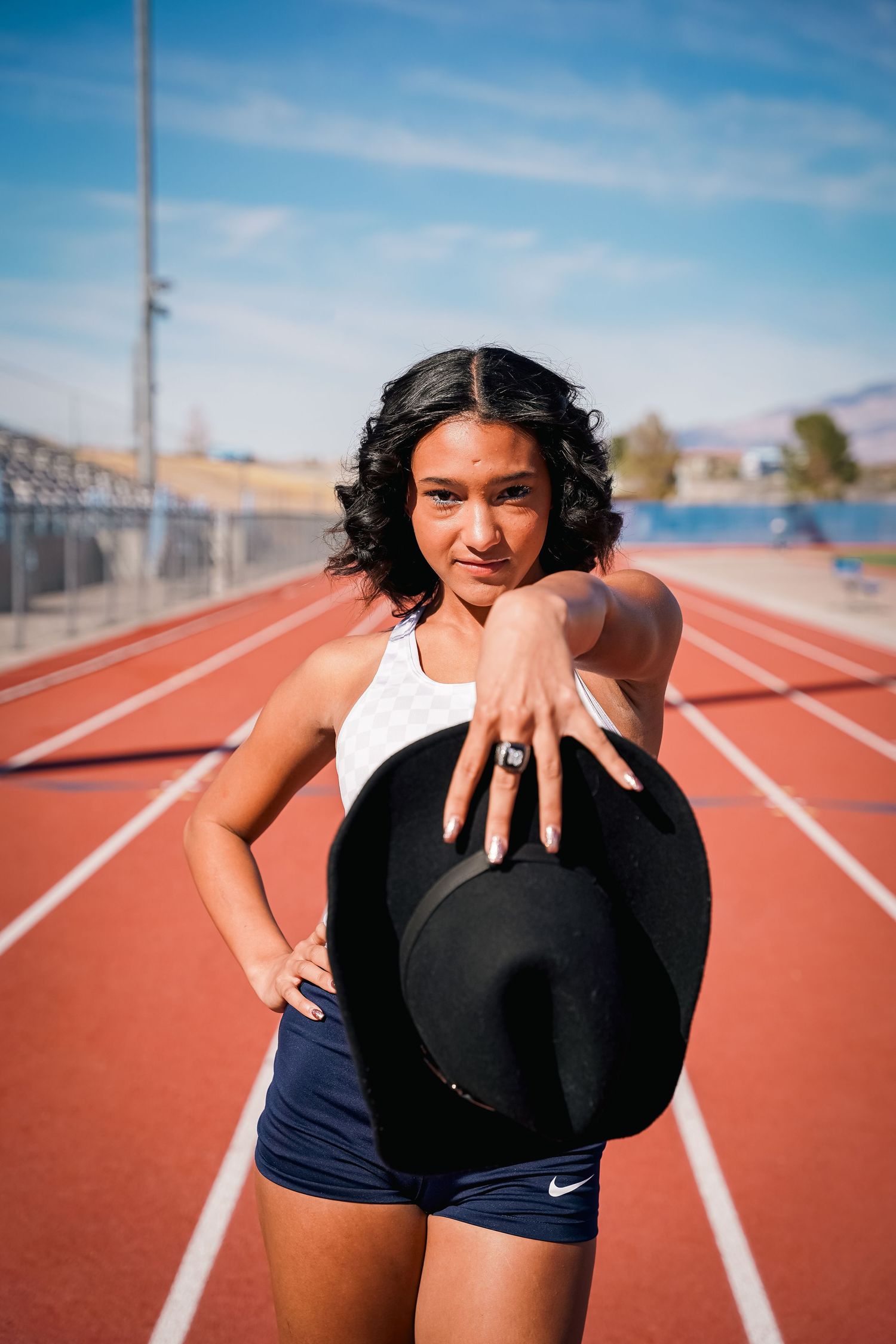 Model poses with black hat on outdoor running track under blue sky.