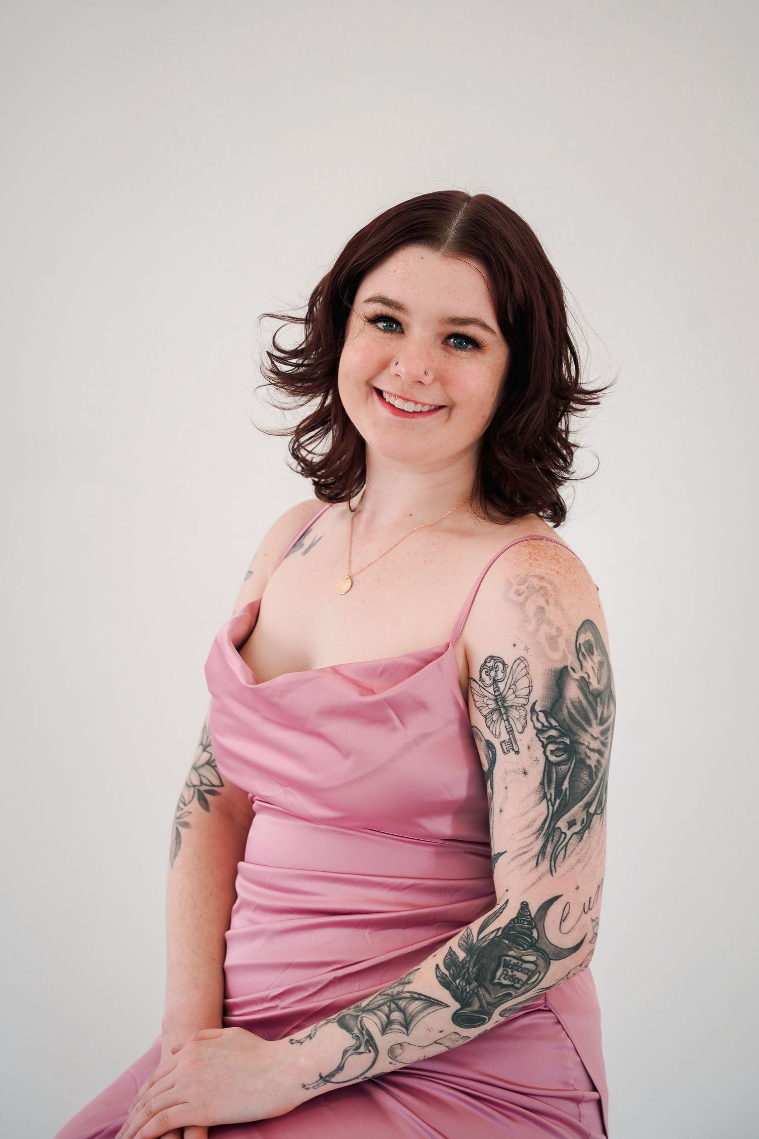 Woman with visible tattoos wearing pink slip dress poses on chair in studio setting.