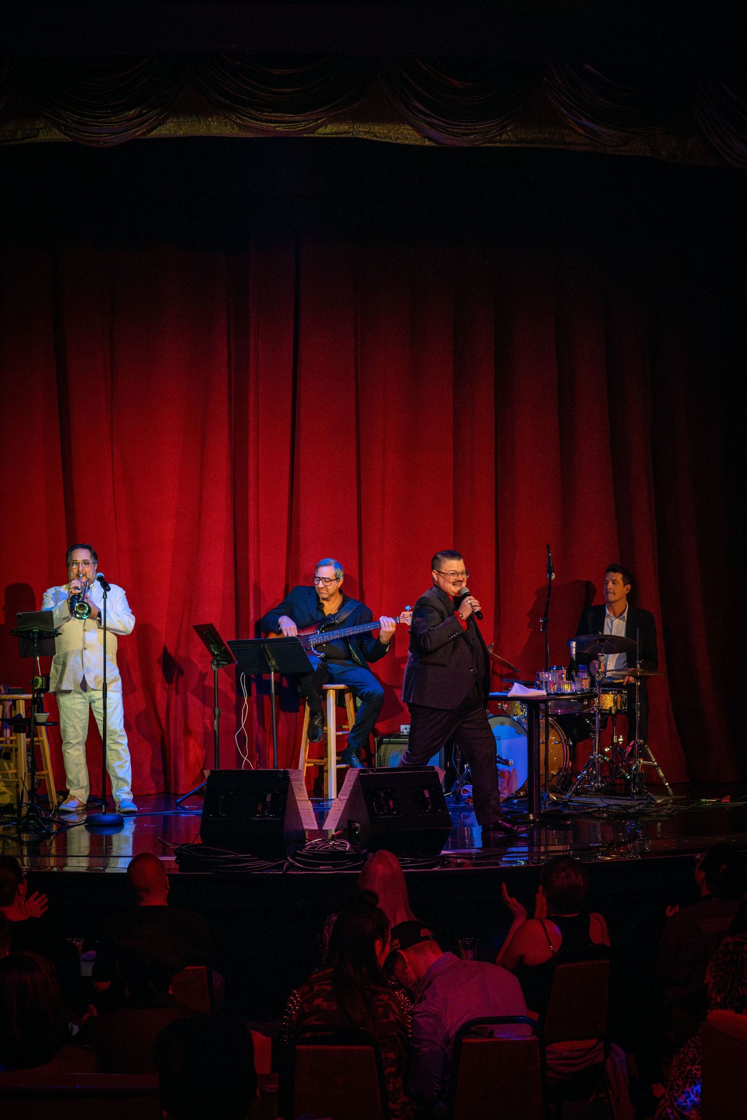 A band performs on stage with red curtains and colorful lighting at a live music venue.