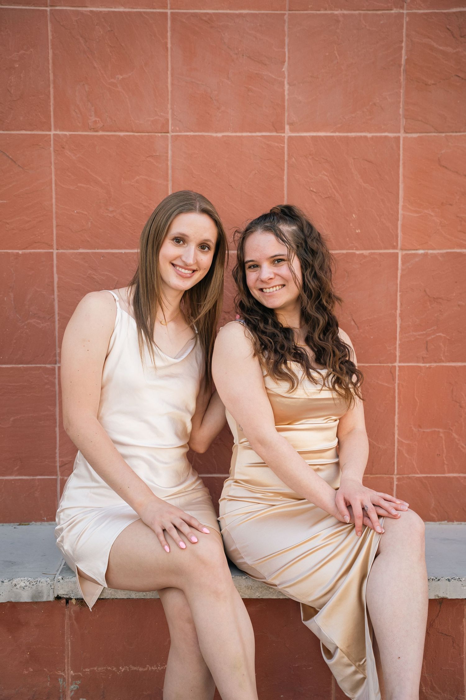 Two women in light colored dresses pose on a bench against a terracotta wall.