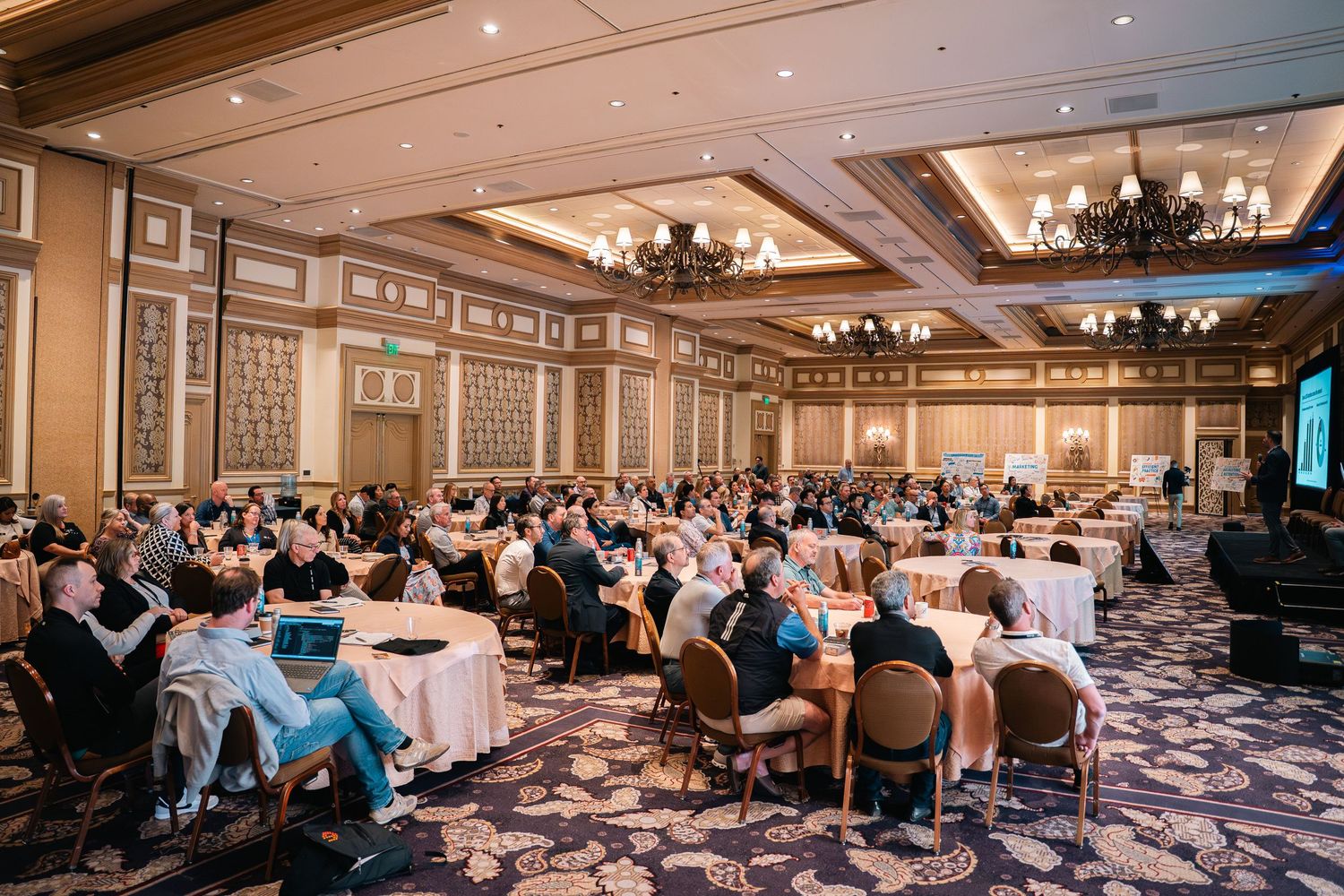 Large conference room filled with attendees at round tables during business event.