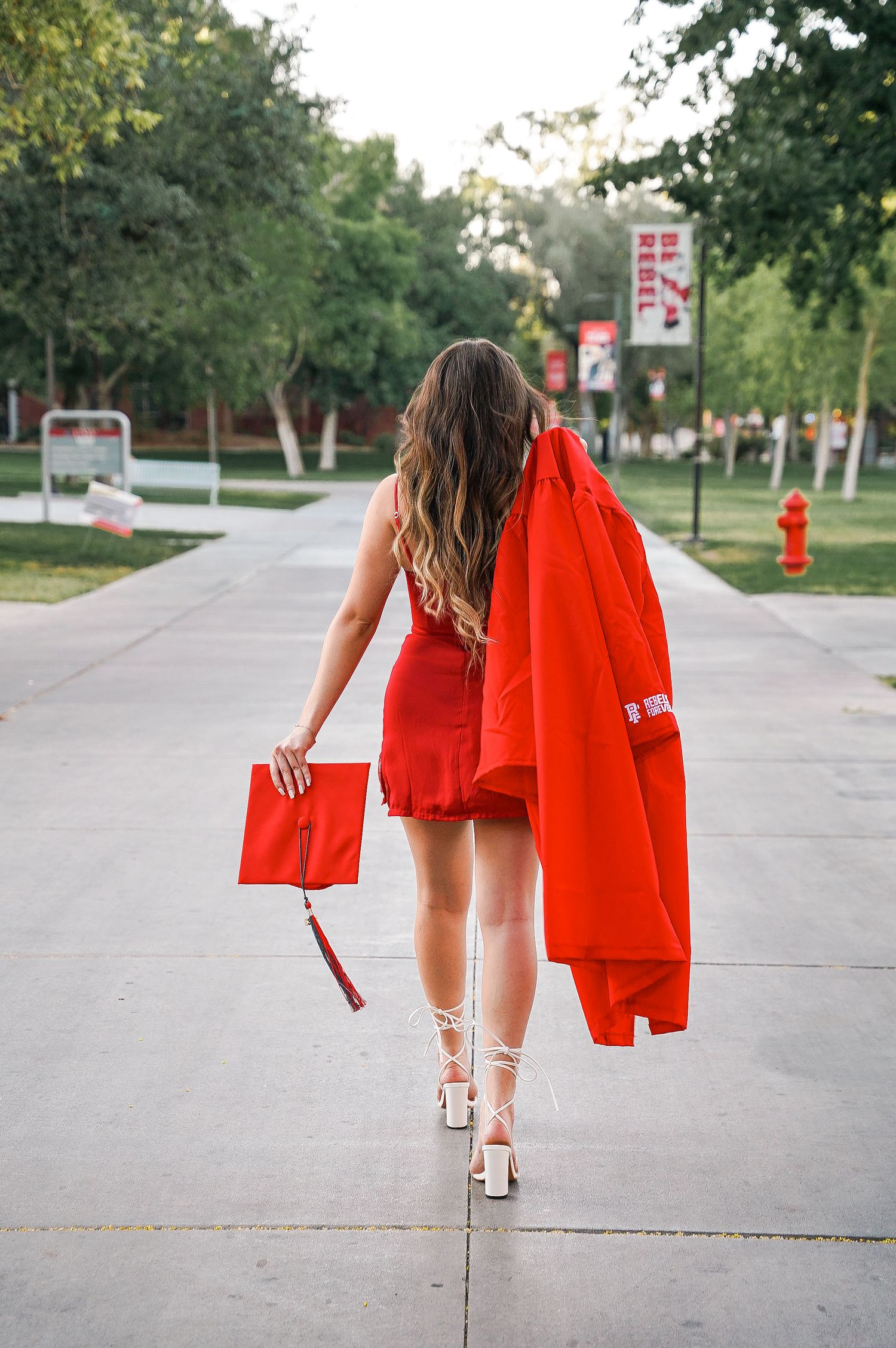 Walking away shot of graduate in red dress carrying cap and gown down campus path.