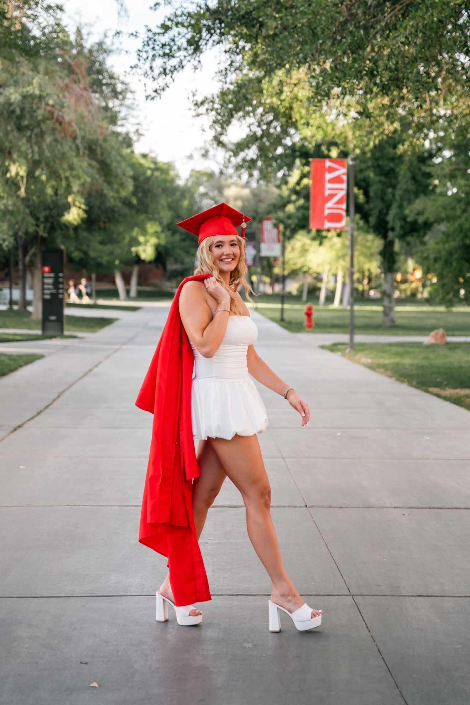 Graduate in red cap and gown poses playfully on campus walkway.