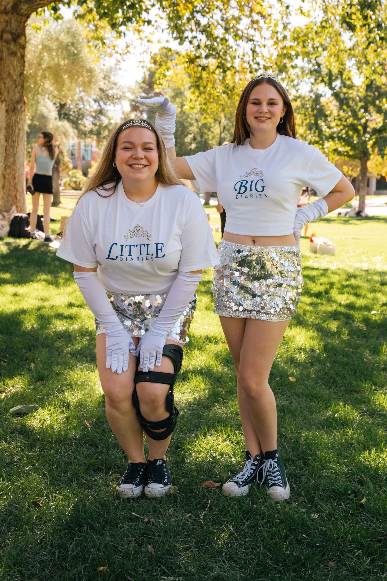 Two friends in matching white shirts and sequin skirts pose on grass wearing 'Big' and 'Little' sorority shirts.