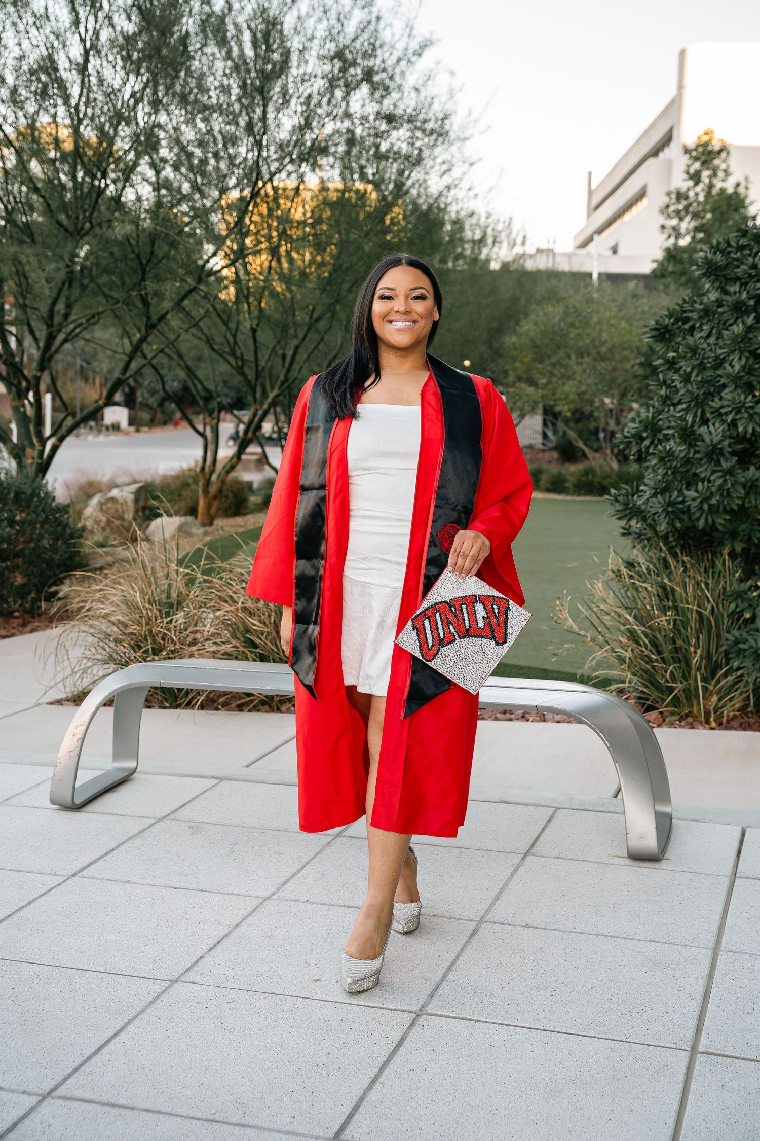 Graduate wearing red gown and white dress posing outdoors with UNLV accessories.